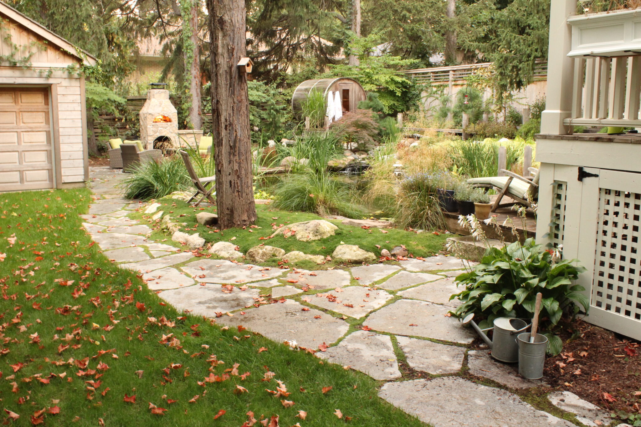 A tranquil backyard with a stone path, birdhouse on a tree, scattered autumn leaves, a pond, and an outdoor fireplace in a lush garden setting.