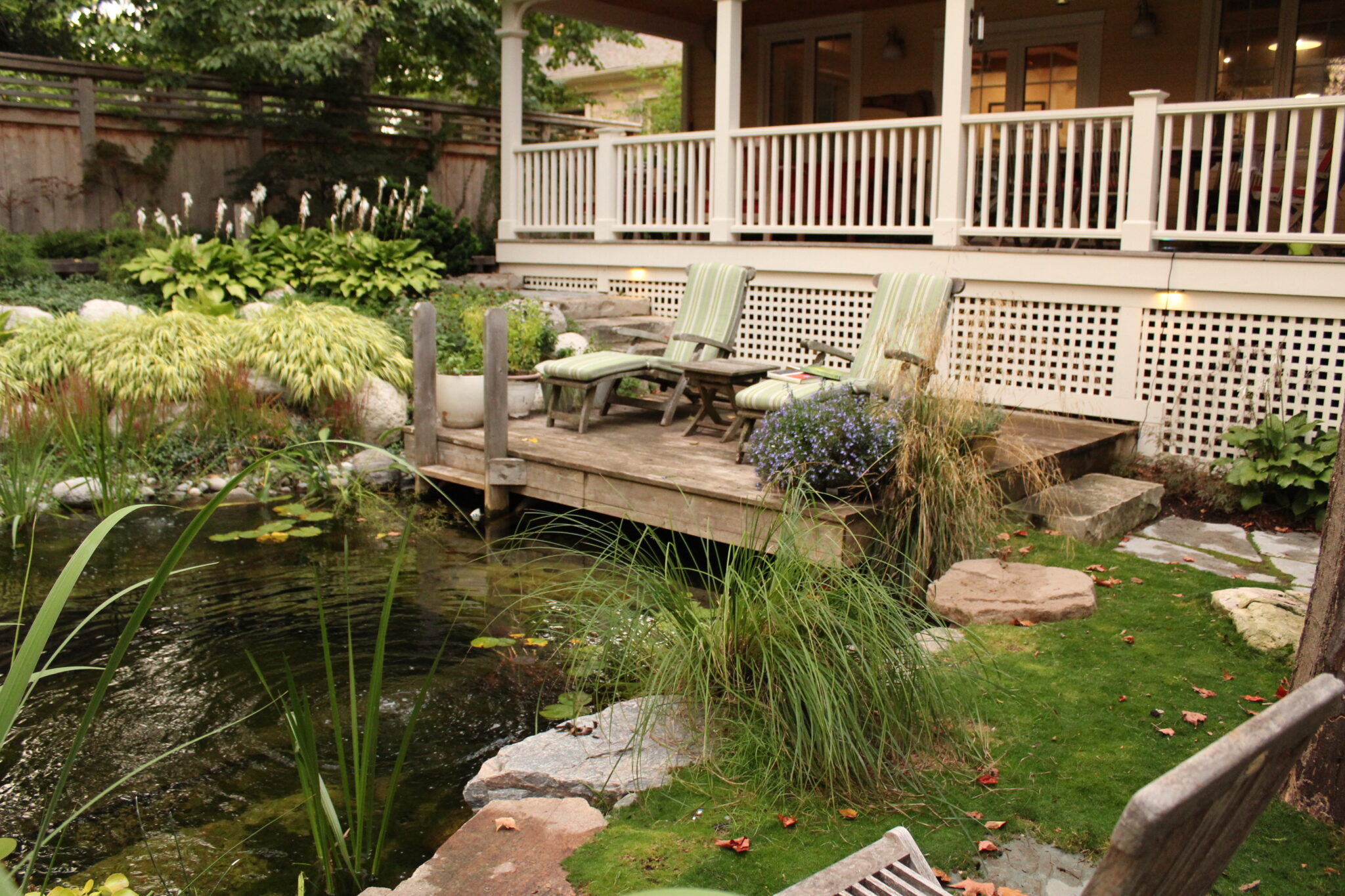 A tranquil backyard setting with a wooden deck, comfortable chairs, lattice privacy screens, and a small serene pond surrounded by lush greenery and stones.