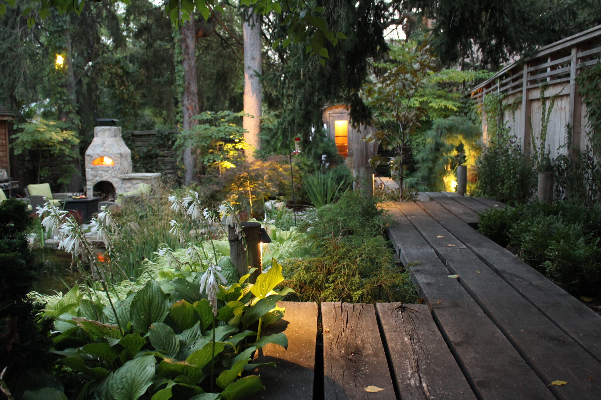 An enchanting garden at dusk with a wood-fired pizza oven, wooden walkway, lush plants, ambient lighting, and a tranquil pond.