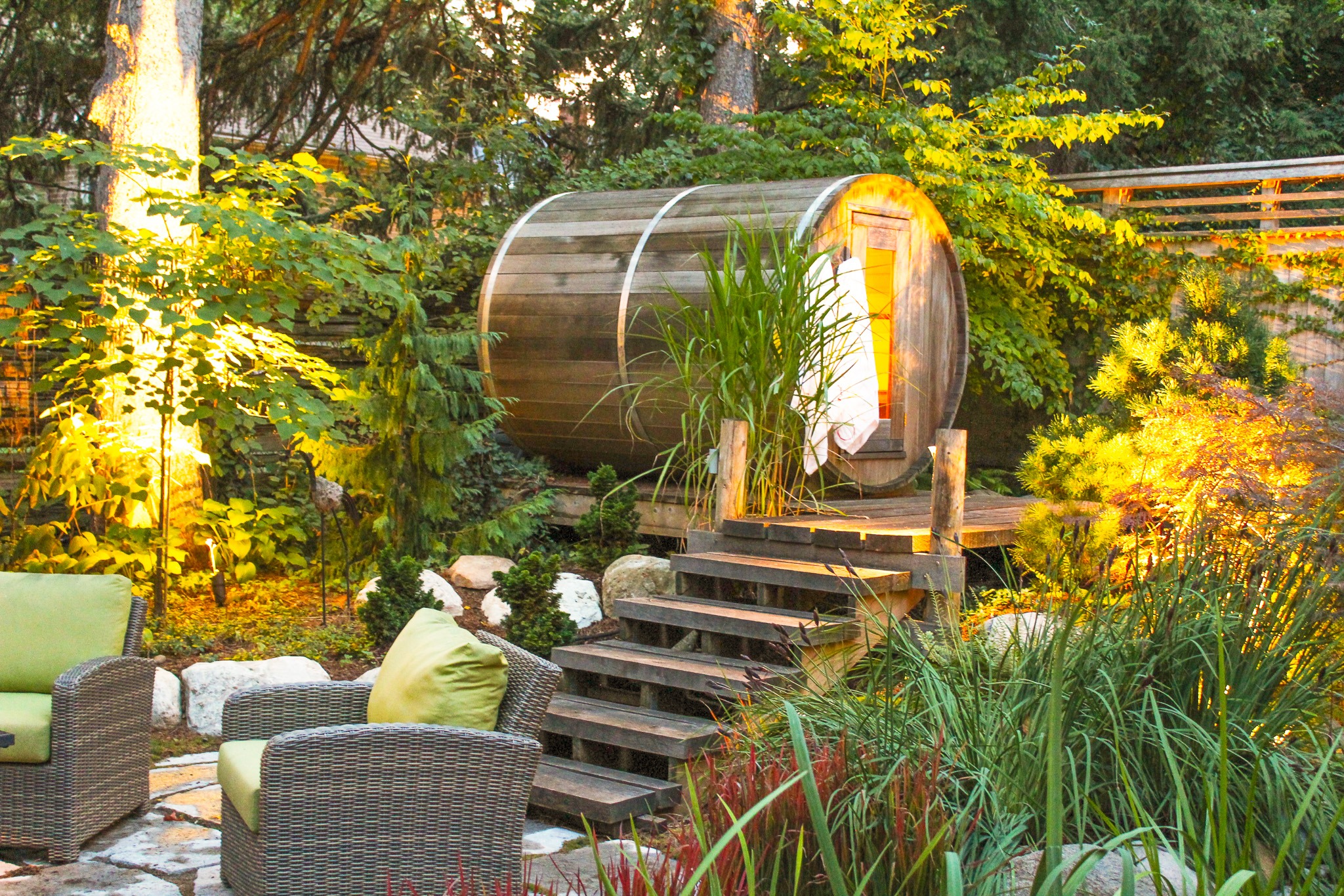 A cozy garden setting features a wooden barrel sauna, comfortable wicker chairs with cushions, lush greenery, and warm lighting at dusk.