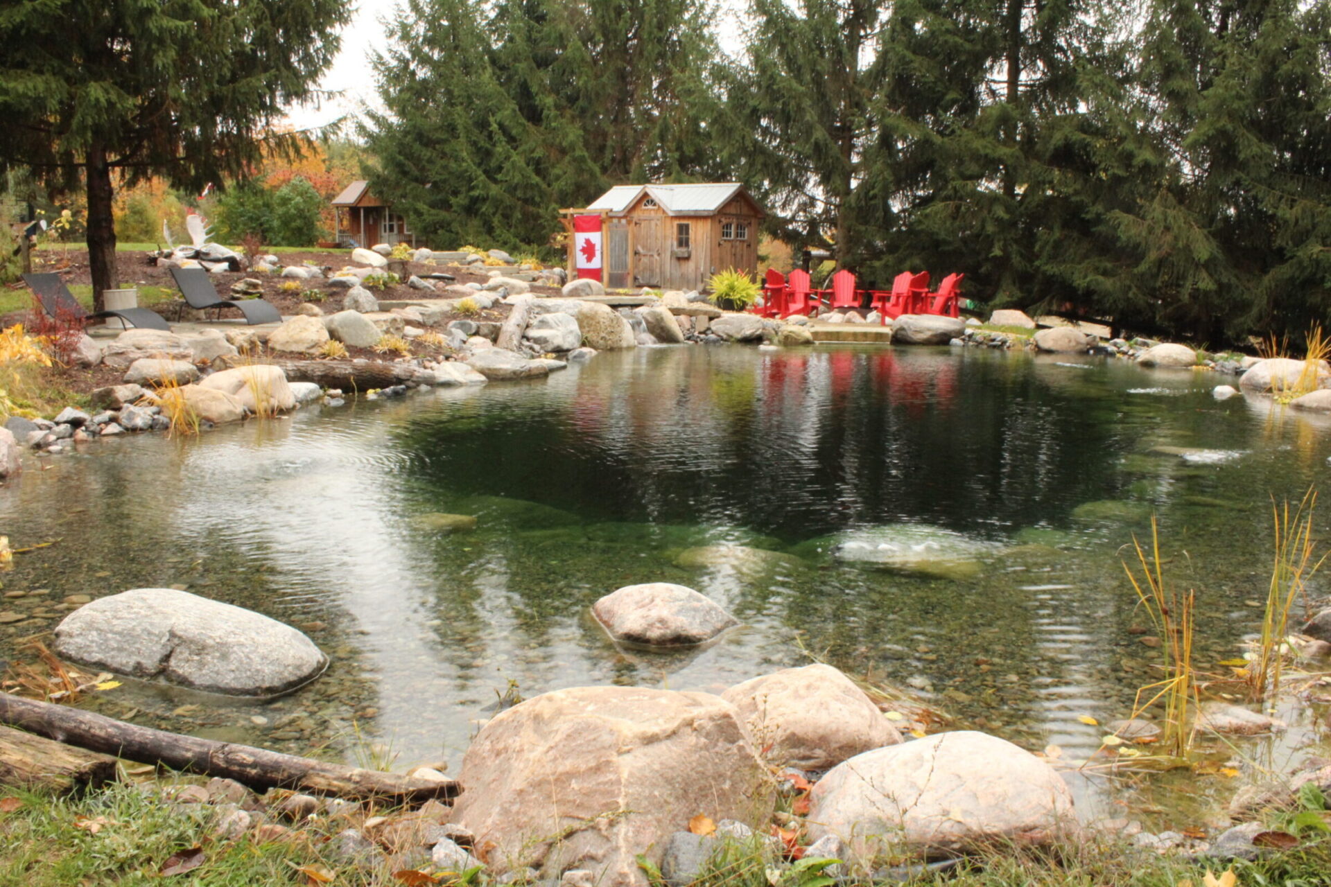 A tranquil pond surrounded by large rocks, flanked by red chairs and coniferous trees, with a cozy wooden shack displaying a Canadian flag.