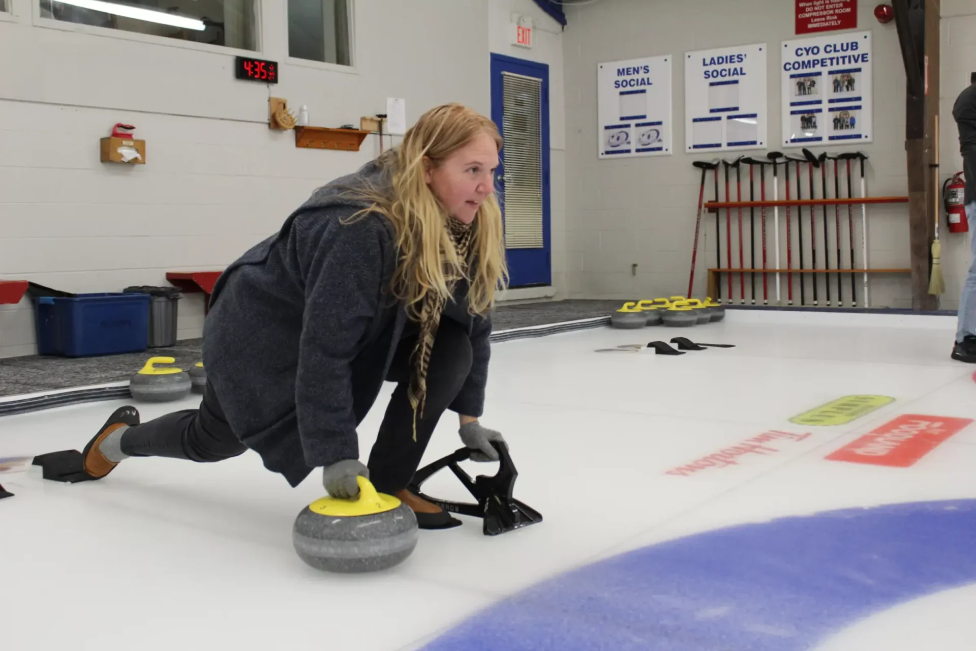 A person is playing curling, sliding on the ice to deliver a stone towards the target, with brooms and game indicators visible in the background.