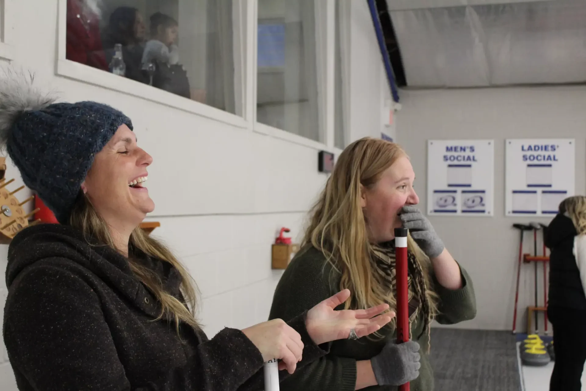 Two people are laughing joyfully inside an indoor ice rink with curling sheets indicated by signs, wearing winter clothes and holding brooms.