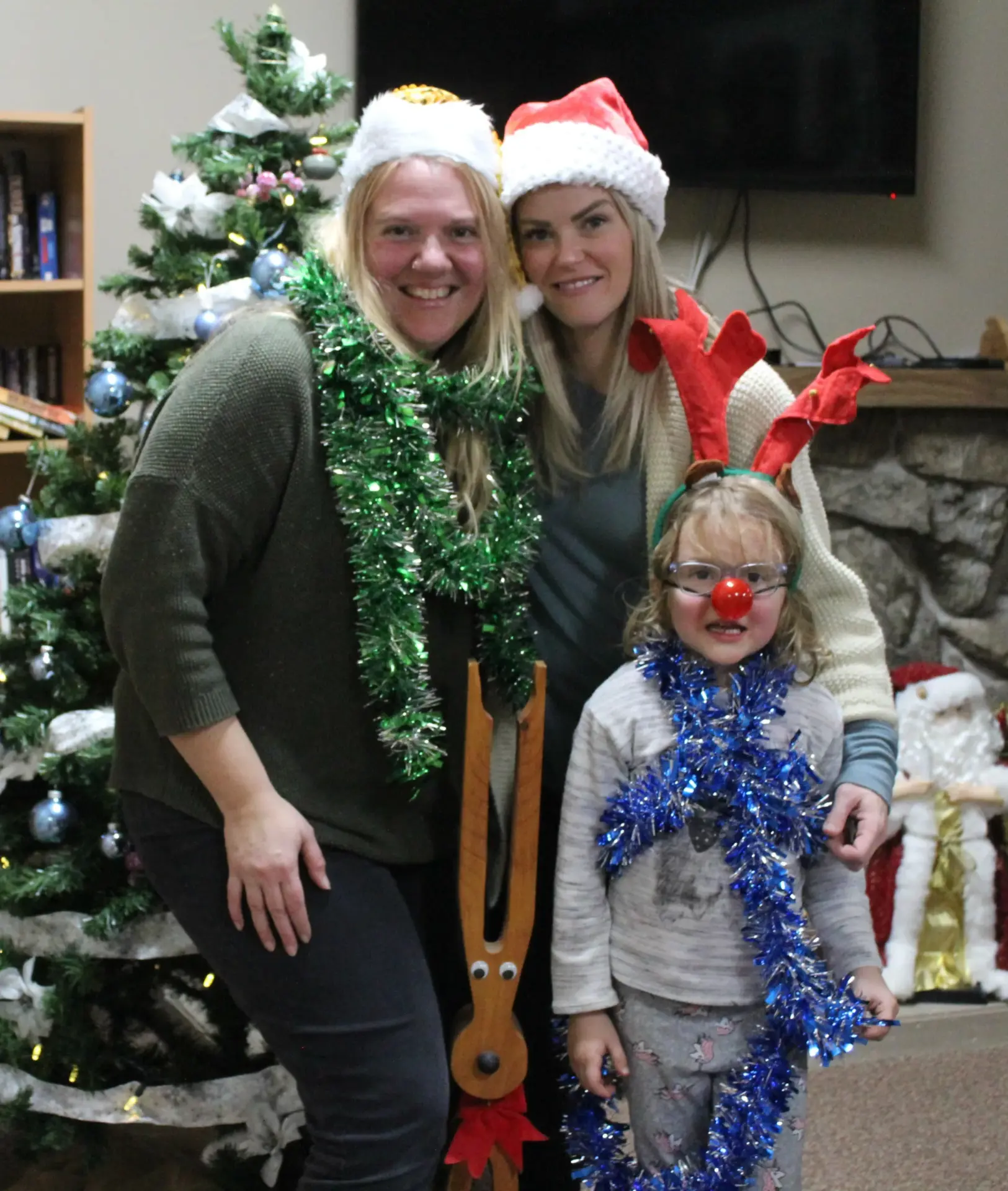 Two adults and a child are wearing festive attire, with Santa hats and tinsel, smiling in front of a decorated Christmas tree and a Santa figure.