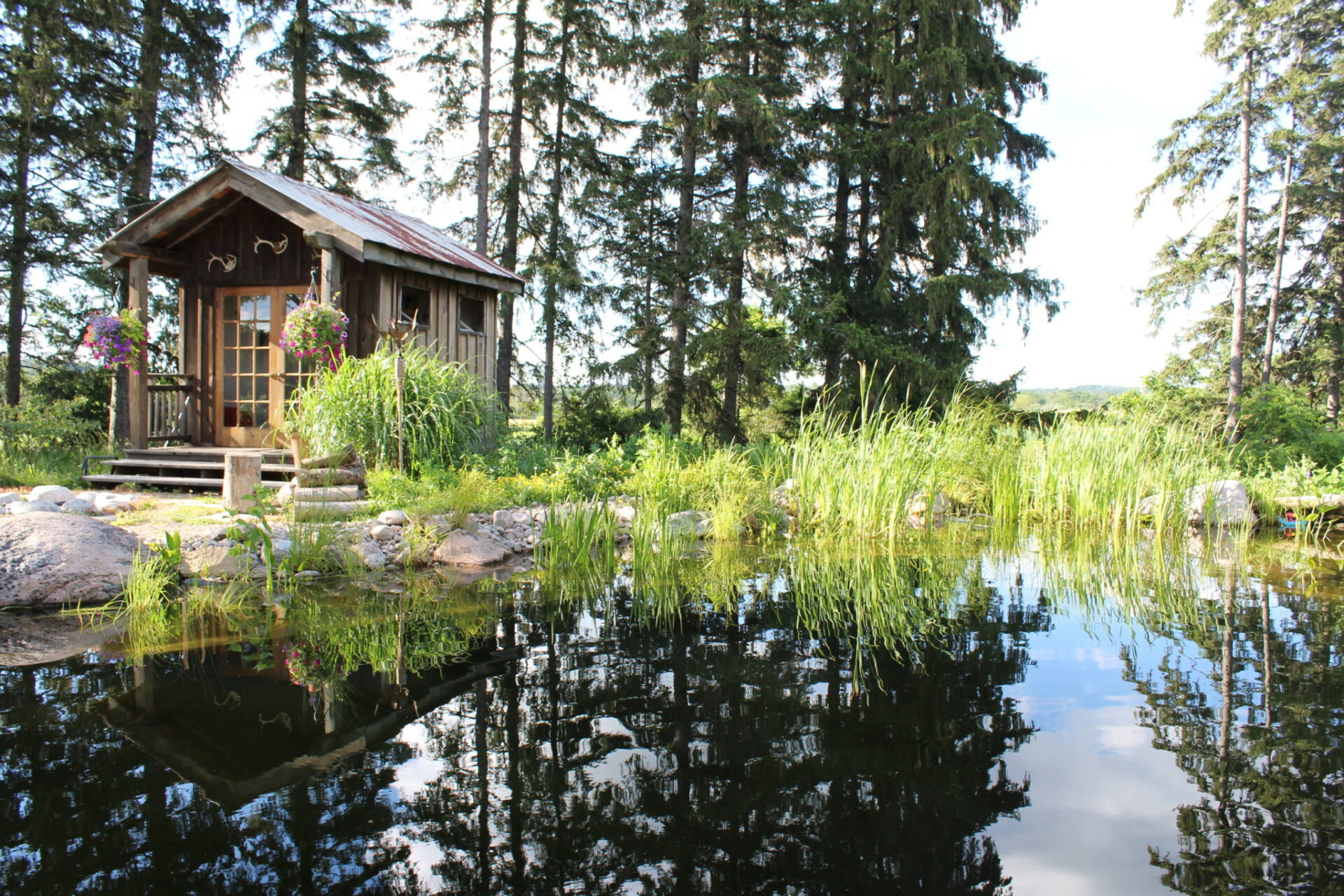 A serene wooden cabin with a porch is surrounded by lush greenery and is reflected in the tranquil water of a pond under a clear blue sky.