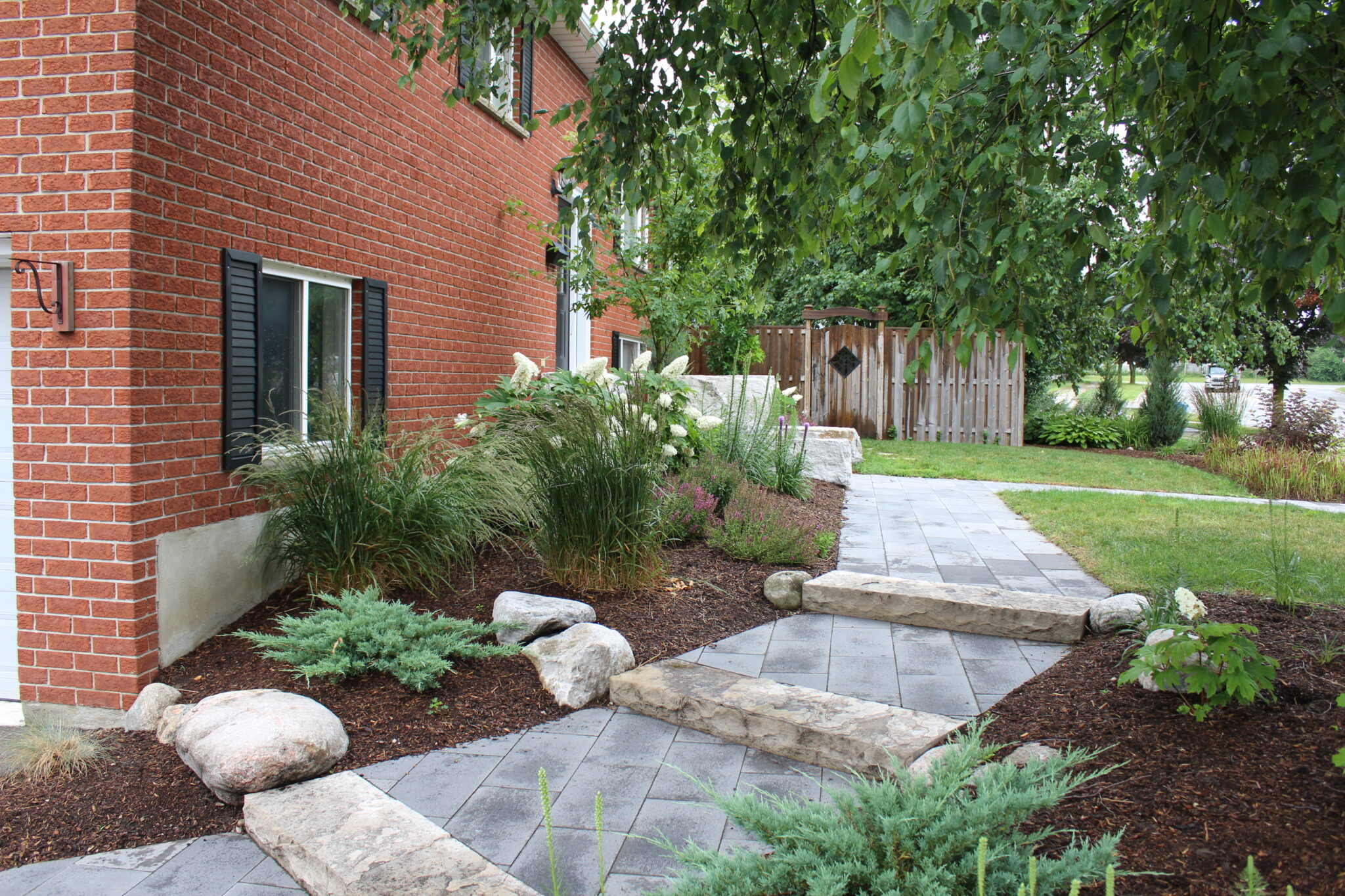 This is a landscaped area beside a brick building featuring a flagstone path, assorted plants, and trees with a wooden fence in the background.