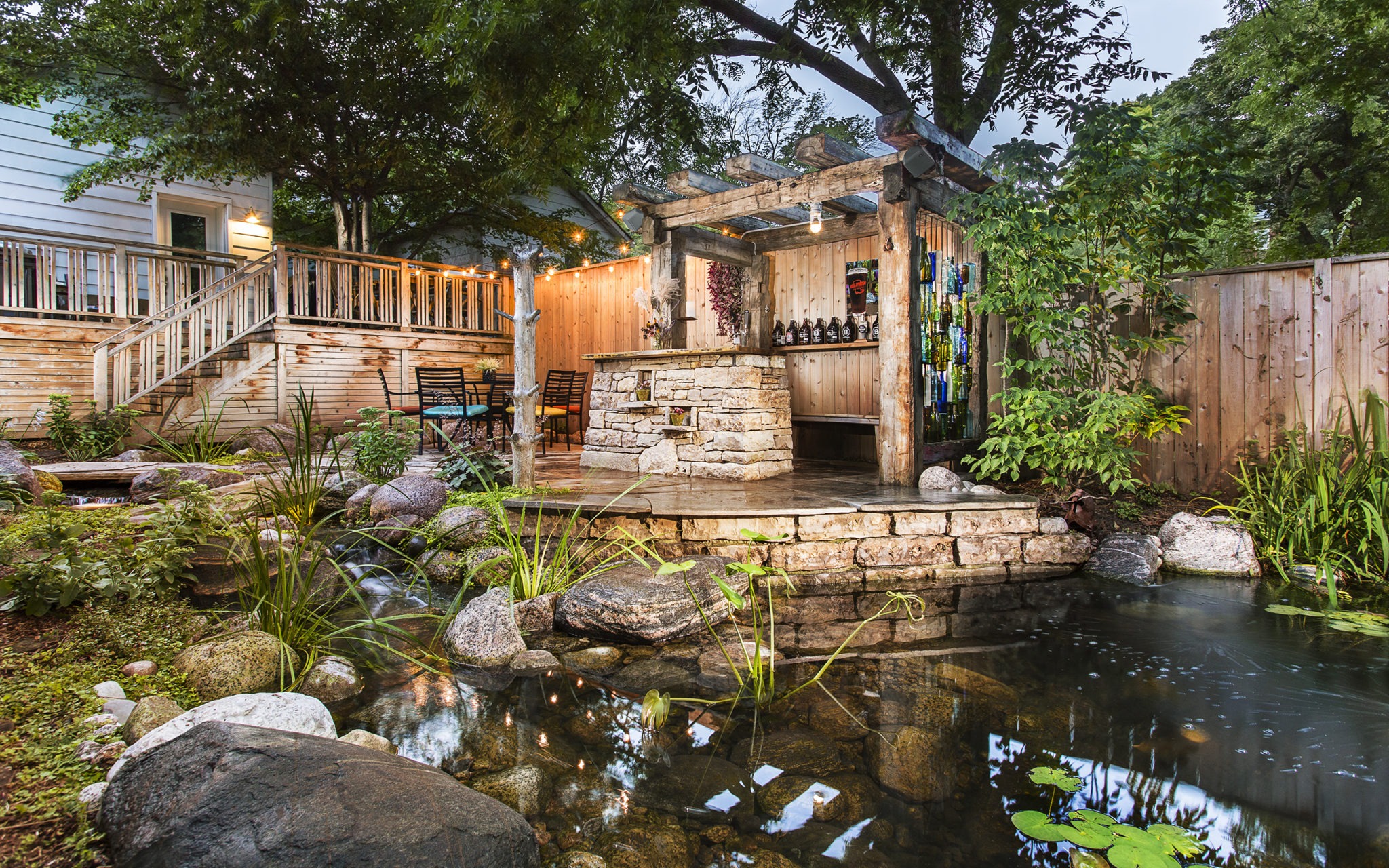 A serene backyard with a pond, lush plants, and a cozy outdoor bar under a pergola, adjacent to a wooden deck leading to a house.