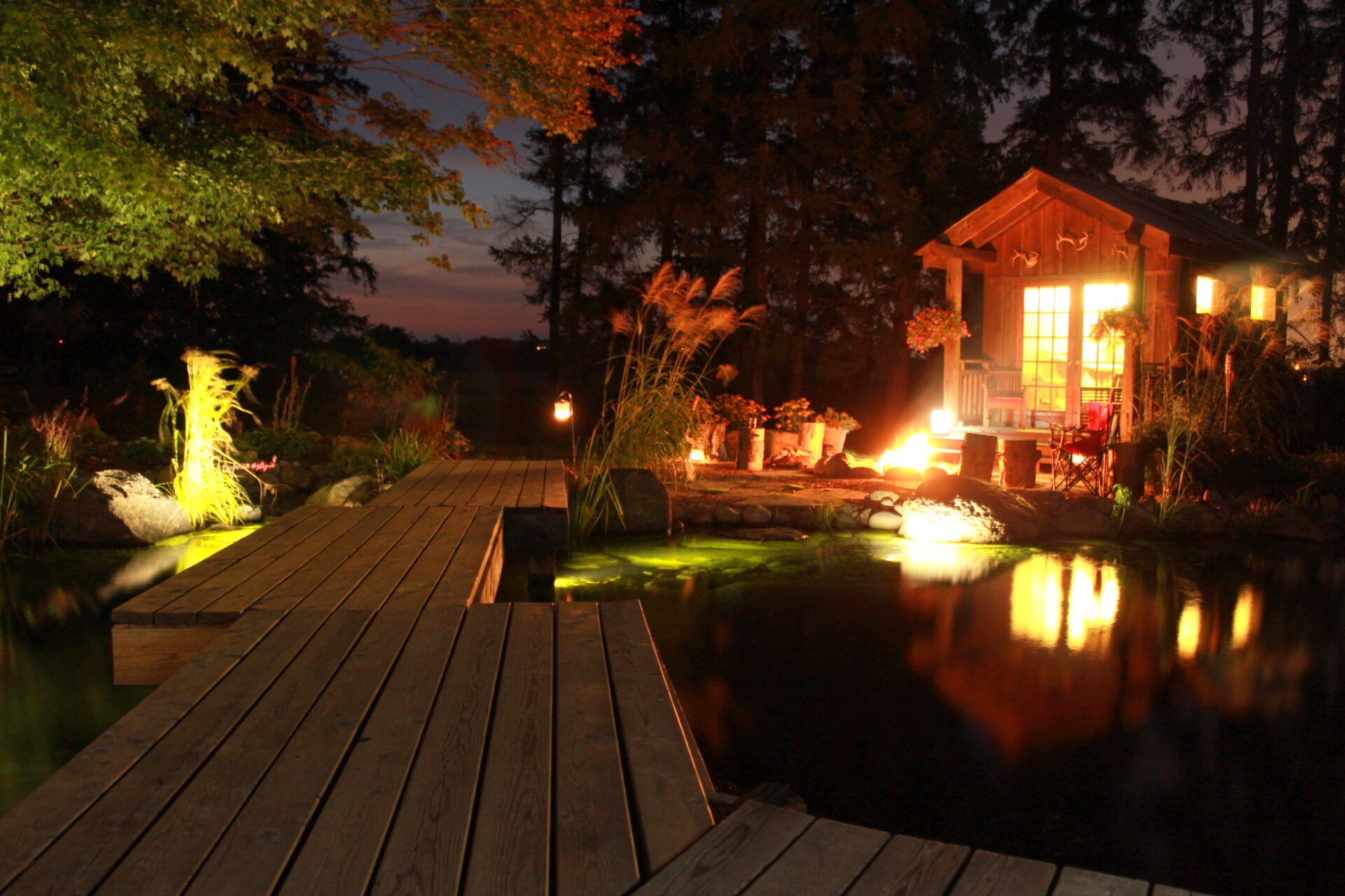 A warmly lit wooden cabin by a pond at dusk, with a dock leading to lush greenery and tranquil waters reflecting the cozy scene.