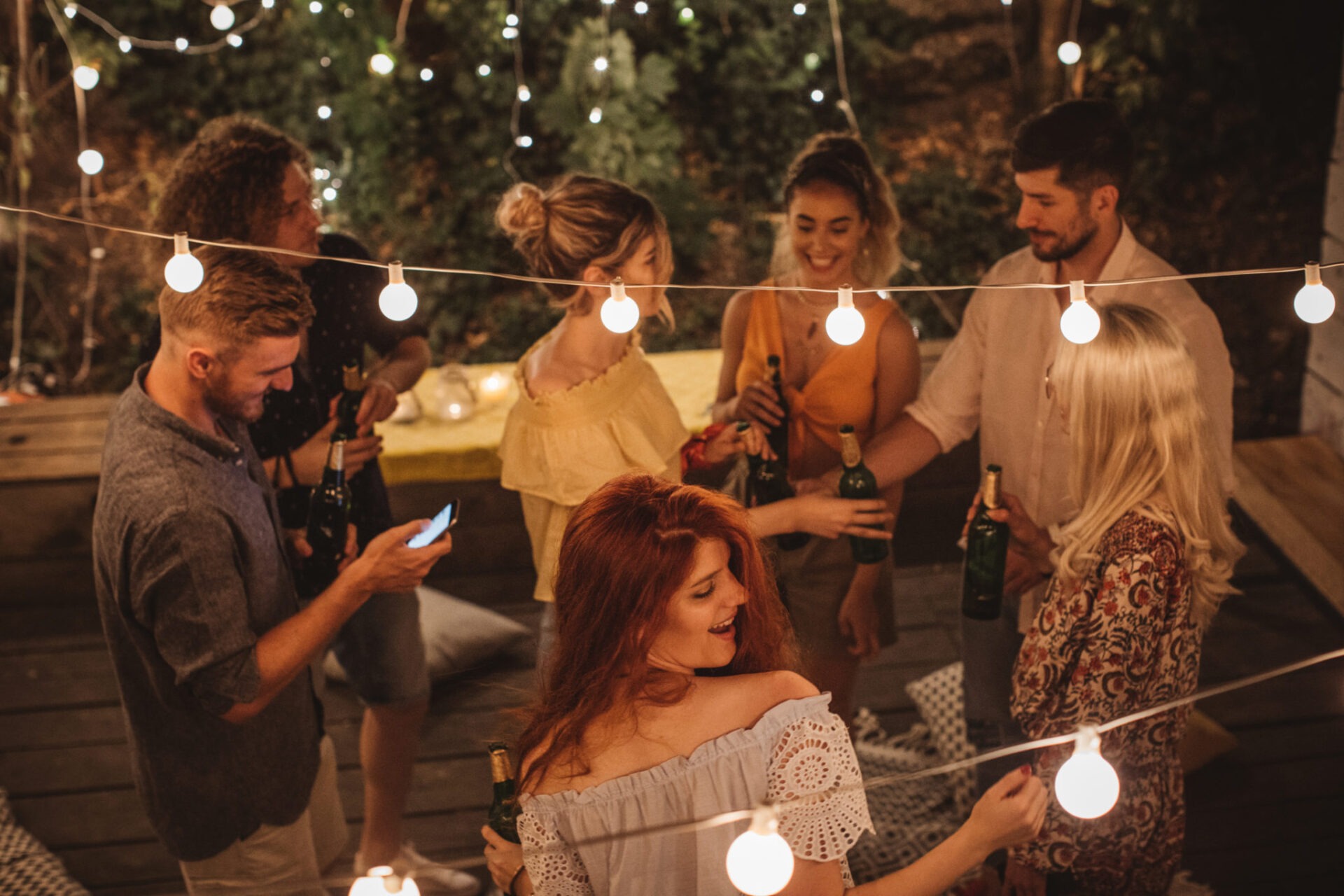 An outdoor gathering at night with string lights, featuring several people chatting and holding drinks, creating a warm, festive atmosphere.