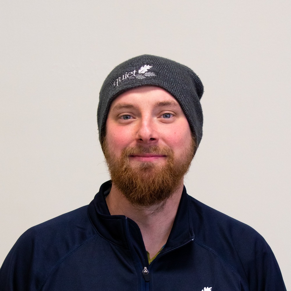 Smiling person with a beard, wearing a knit hat and zip-up top, looking at the camera. Background is plain and light-colored.