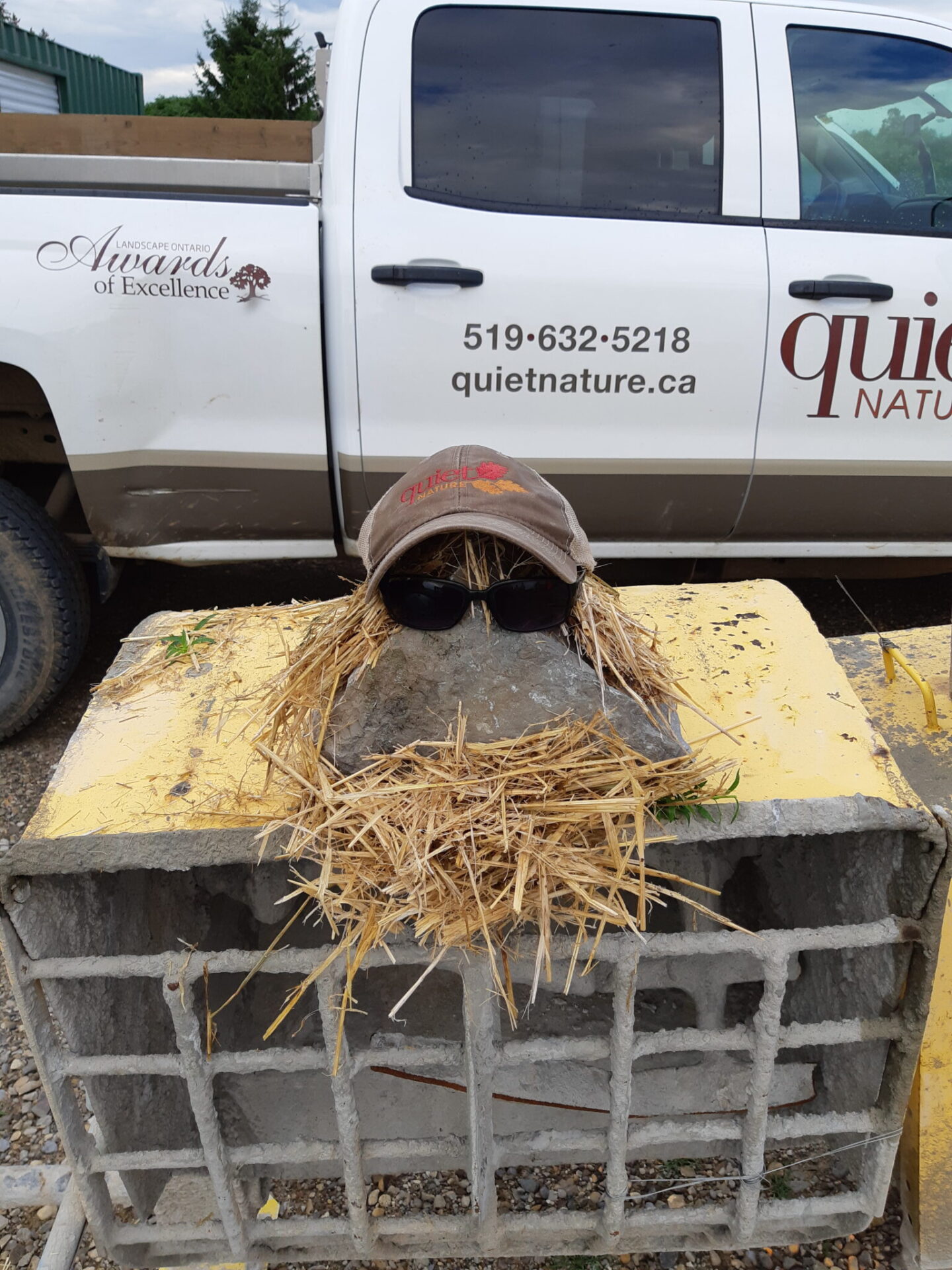 A concrete block with straw, sunglasses, and a hat, resembling a face, is in front of a white truck with company details on the door.