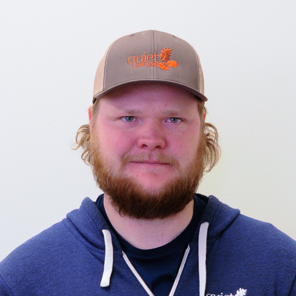 A person with a brown beard is wearing a brown cap and a blue hoodie, standing against a white background, looking at the camera.