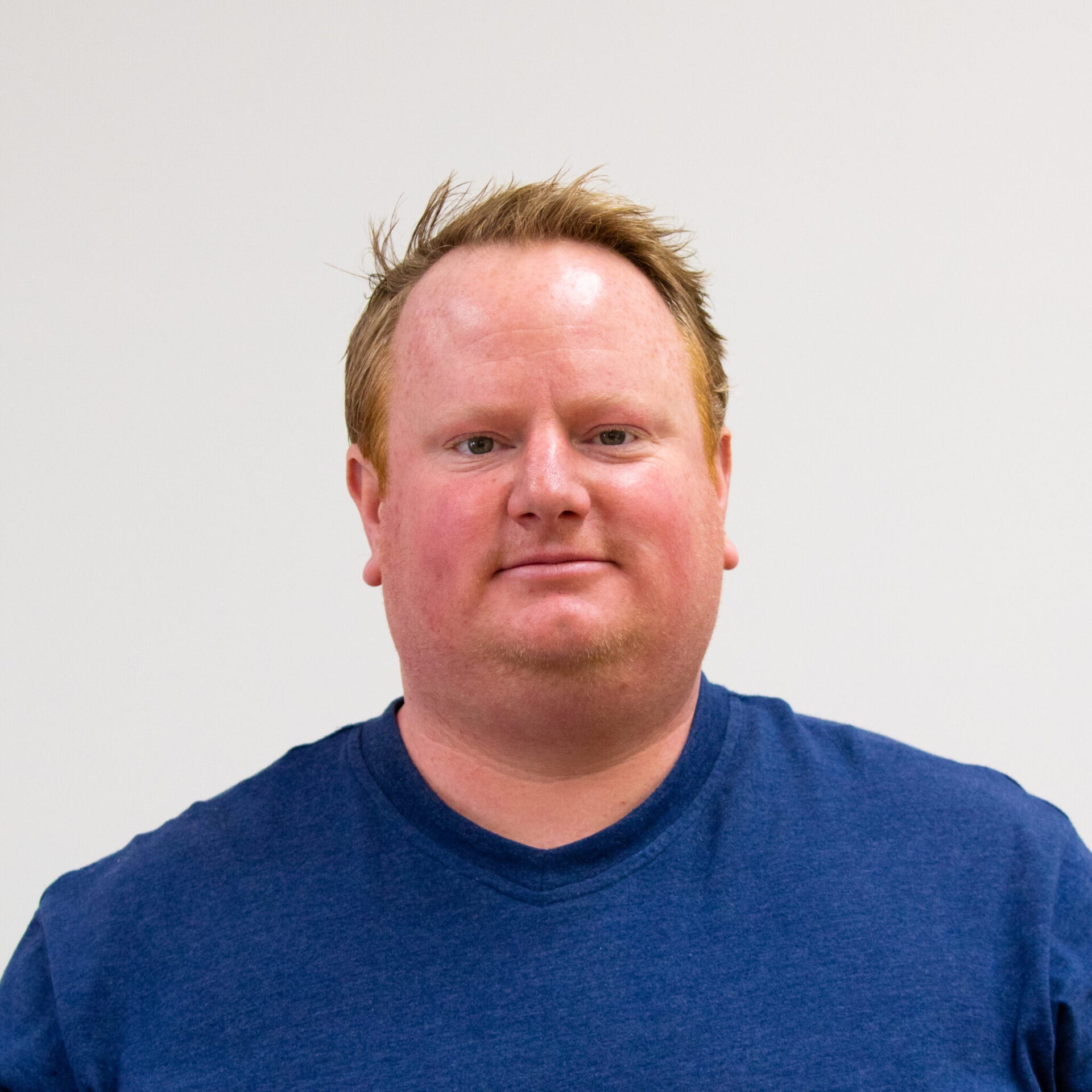 The image shows a person with short spiked hair, wearing a blue crew neck t-shirt, against a neutral background, looking directly at the camera.