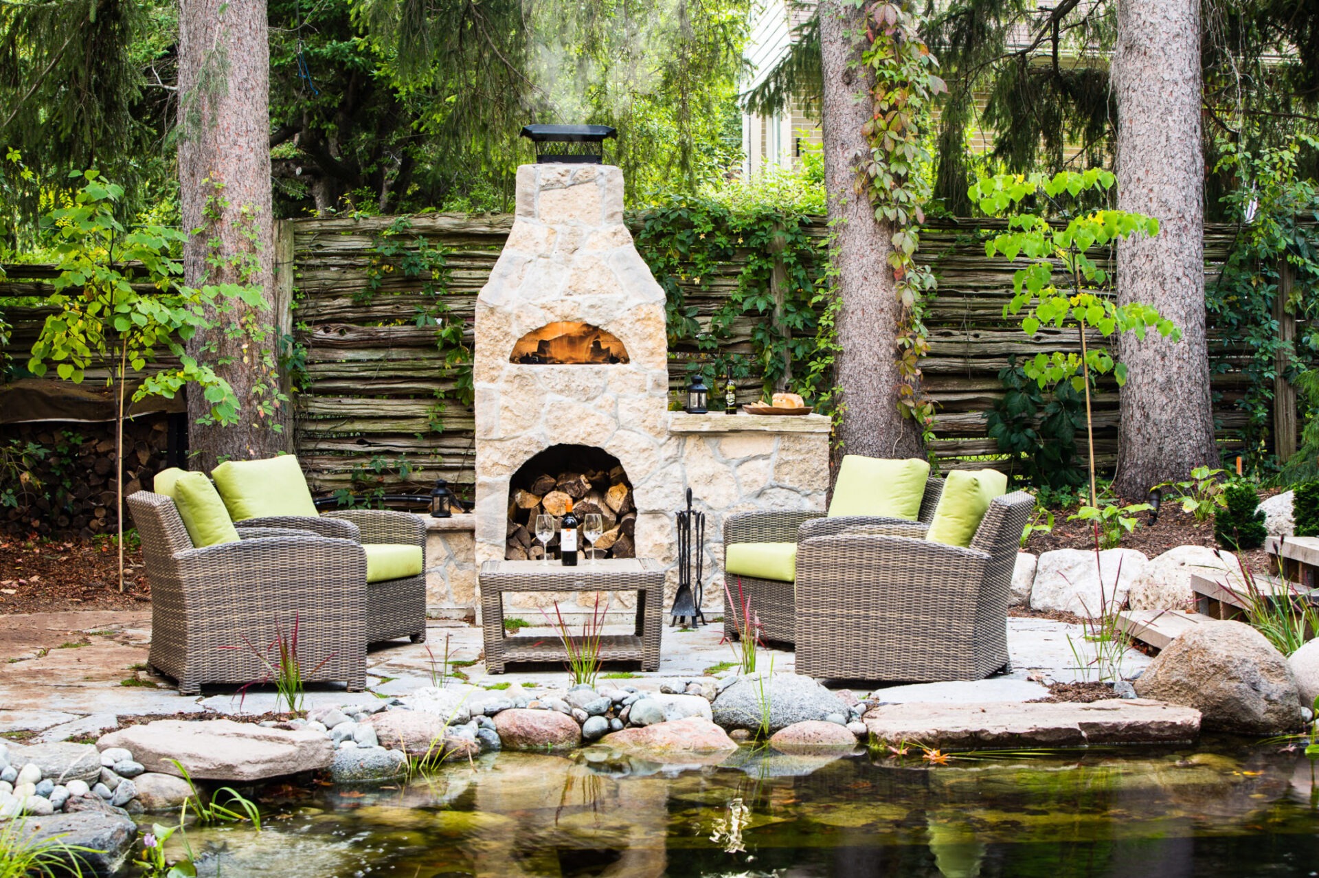 An outdoor patio scene with wicker chairs, green cushions, a stone fireplace, and a pond, surrounded by trees and a wooden log wall.