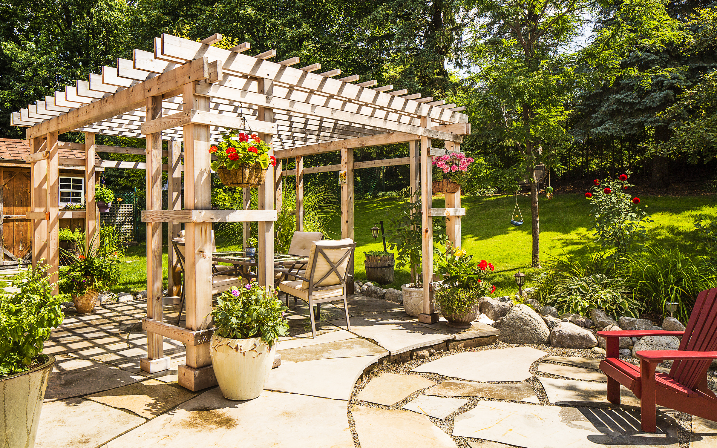 image of a backyard with a large wooden frame terrace decorated with hanging planters. the terrace is on a patio of large flat tiles of uniquely cut pale rock slabs.