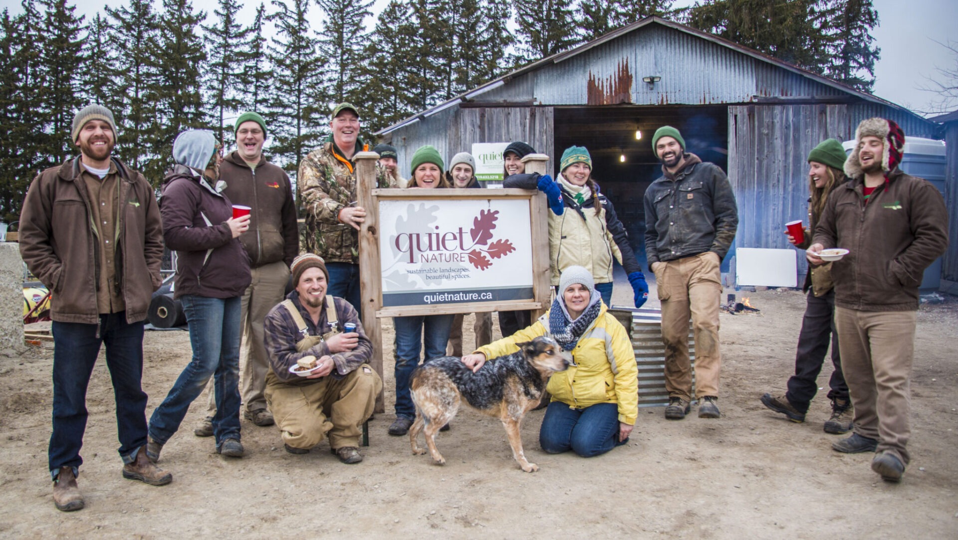 A group of people stands outdoors holding a sign with "Quiet Nature" and a URL on it. A dog is present. Smiling faces suggest a casual, happy gathering.