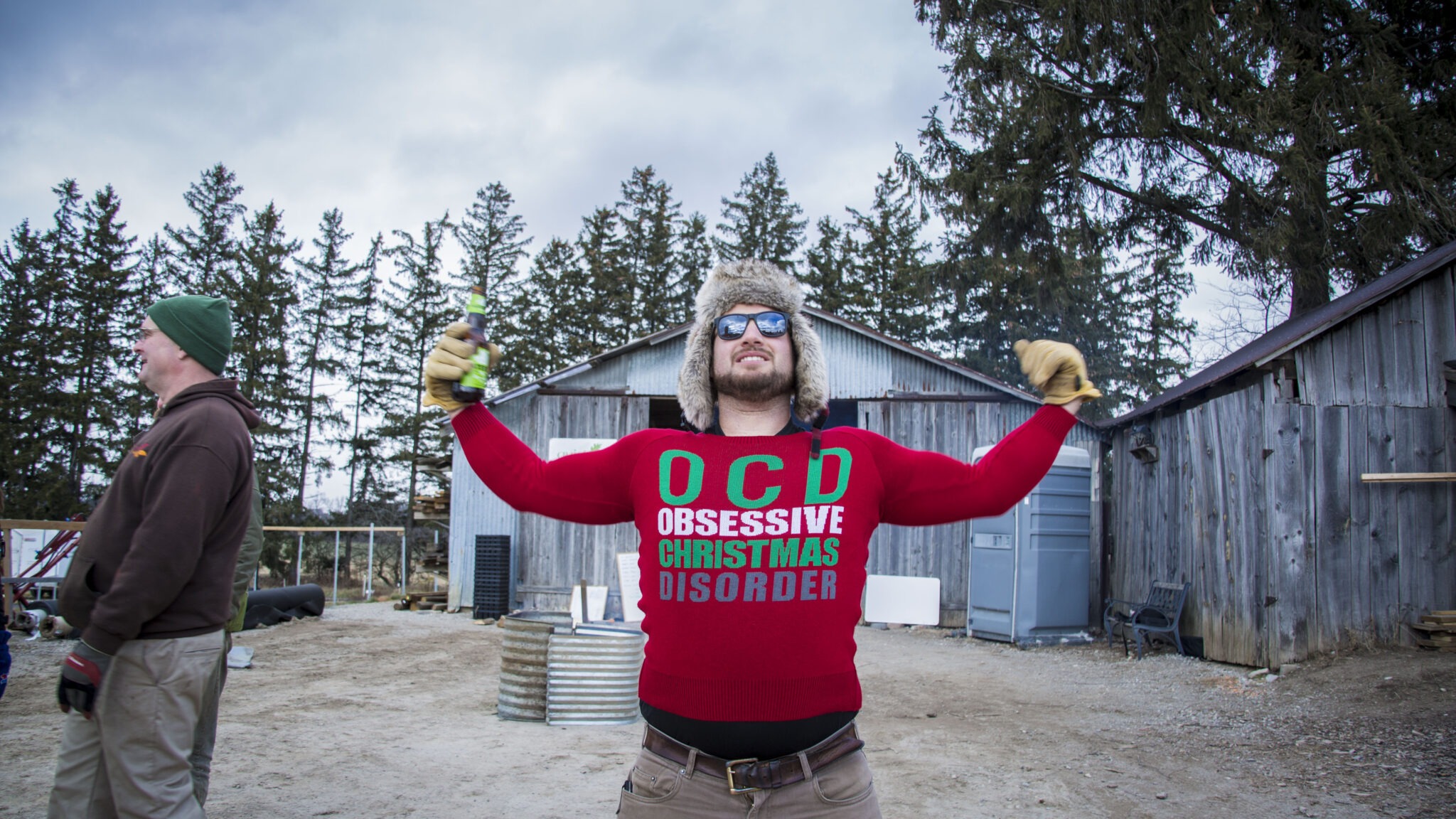 A person in a red sweater with "OCD - Obsessive Christmas Disorder" stands triumphantly in a rural setting, another person in the background smiles.