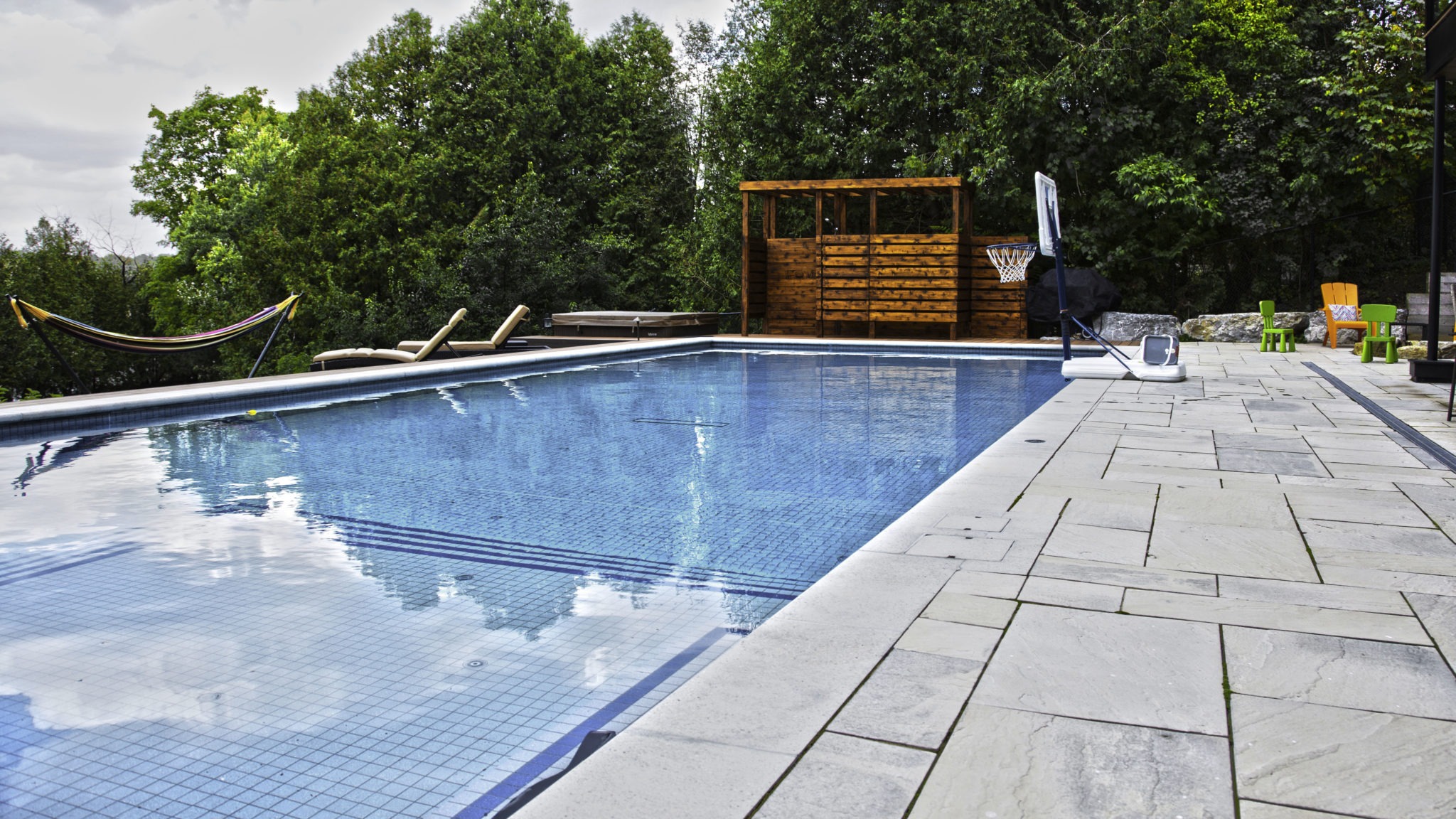 A serene outdoor pool surrounded by trees features sun loungers, a hammock, a basketball hoop, and colorful chairs on a paved deck.