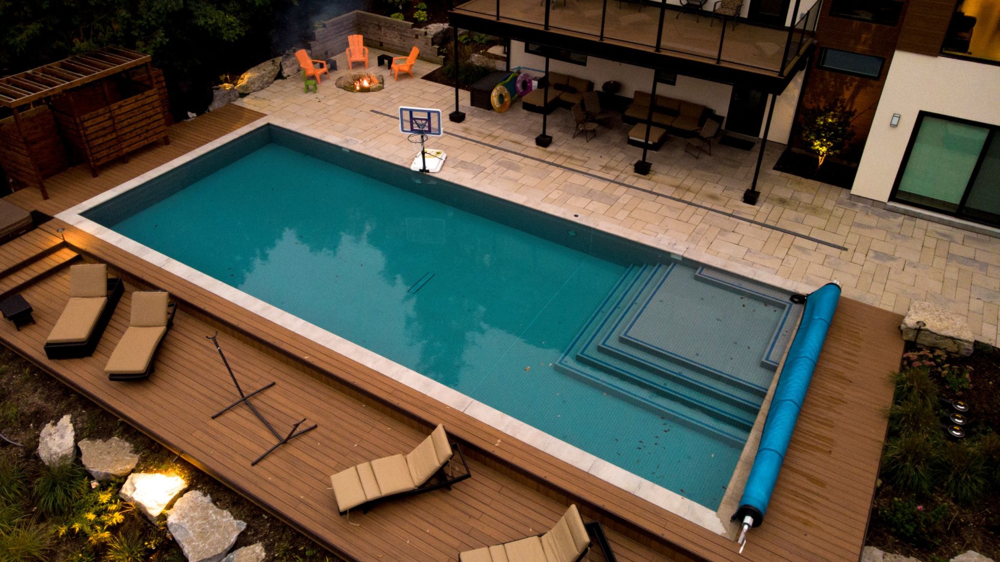 An aerial view of a backyard with a rectangular pool, wooden deck, sun loungers, a basketball hoop, and an outdoor patio area with a fire pit.