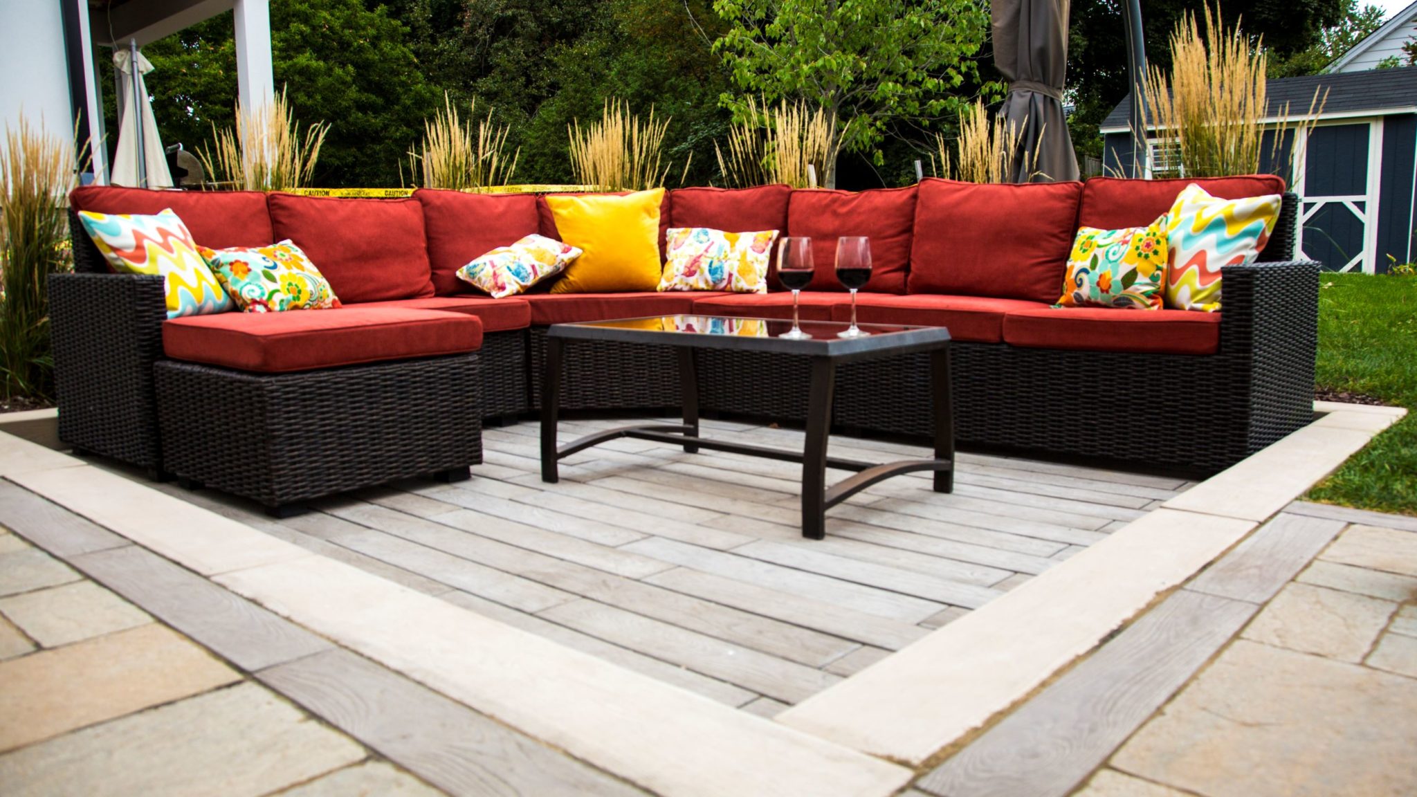 An outdoor seating area with a red cushioned wicker sofa, colorful pillows, a glass-top coffee table, two wine glasses, and surrounding greenery.