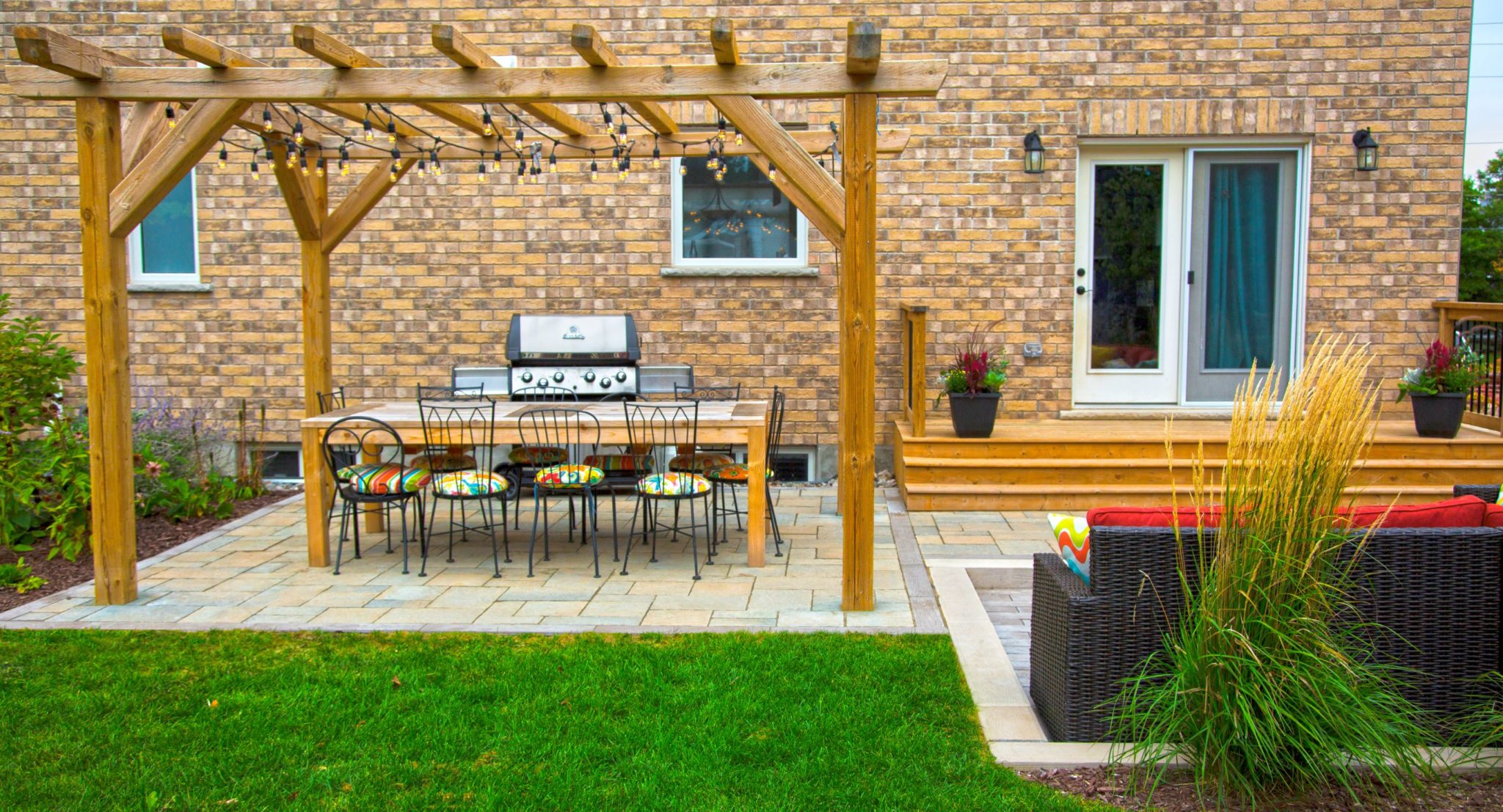 A cozy backyard with a wooden pergola, outdoor dining set, grill, lounging area, green lawn, and landscaped garden beside a brick house.