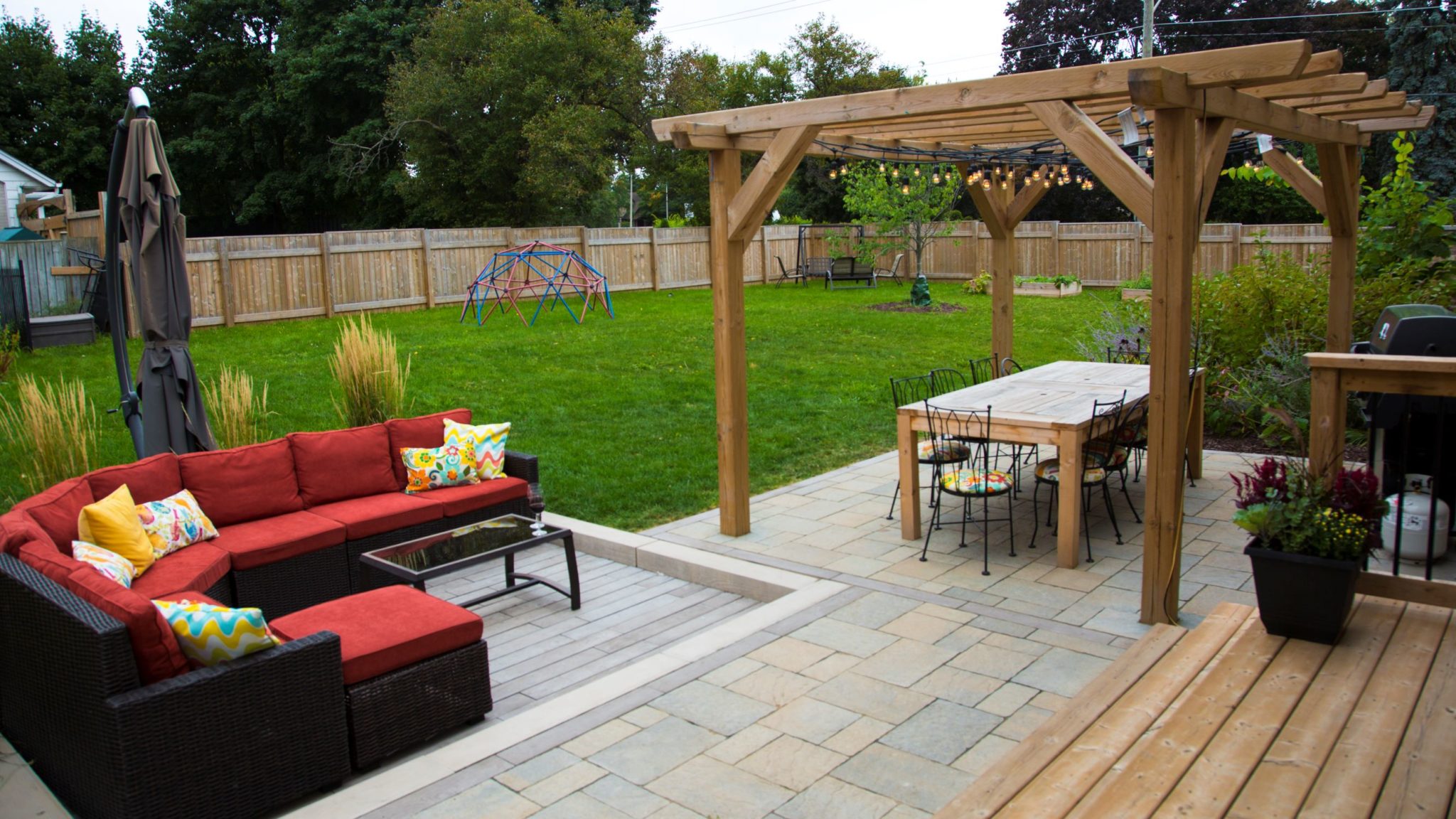 A cozy backyard with a red sofa set, a wooden pergola, string lights, a dining area, a playground, a lush lawn, and a privacy fence.