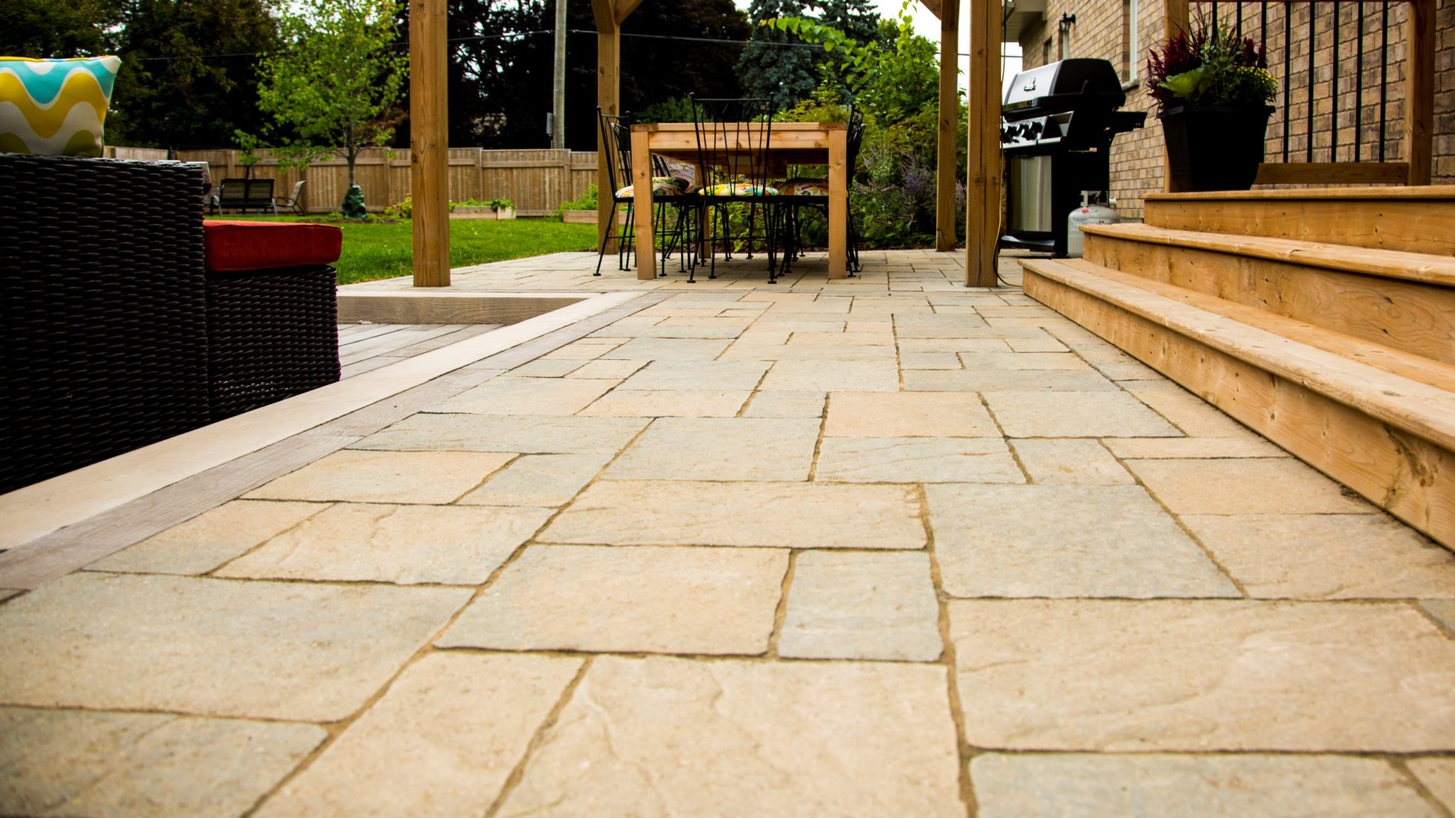 A patio with large stone tiles, wooden steps leading to a house, outdoor furniture, a grill, and a lush garden visible in the background.