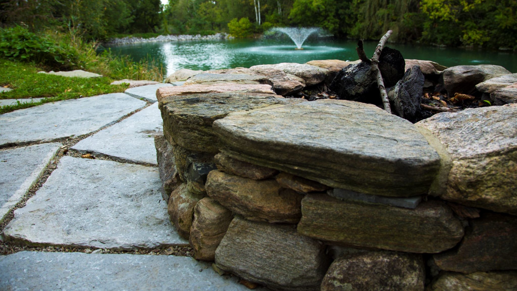 A stone fire pit with charred wood sits beside a flagstone path with a tranquil pond and fountain in the background, surrounded by greenery.