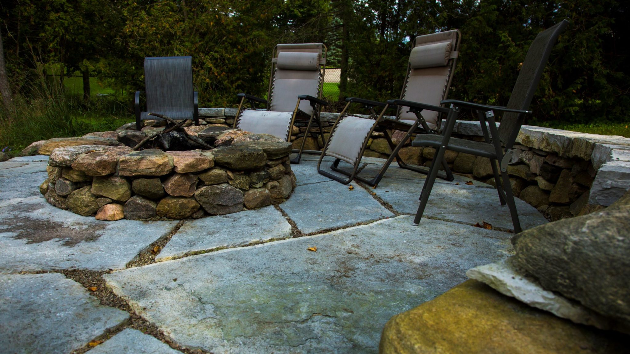 An outdoor stone fire pit surrounded by patio chairs on a flagstone terrace. The setting appears tranquil with green vegetation in the background.