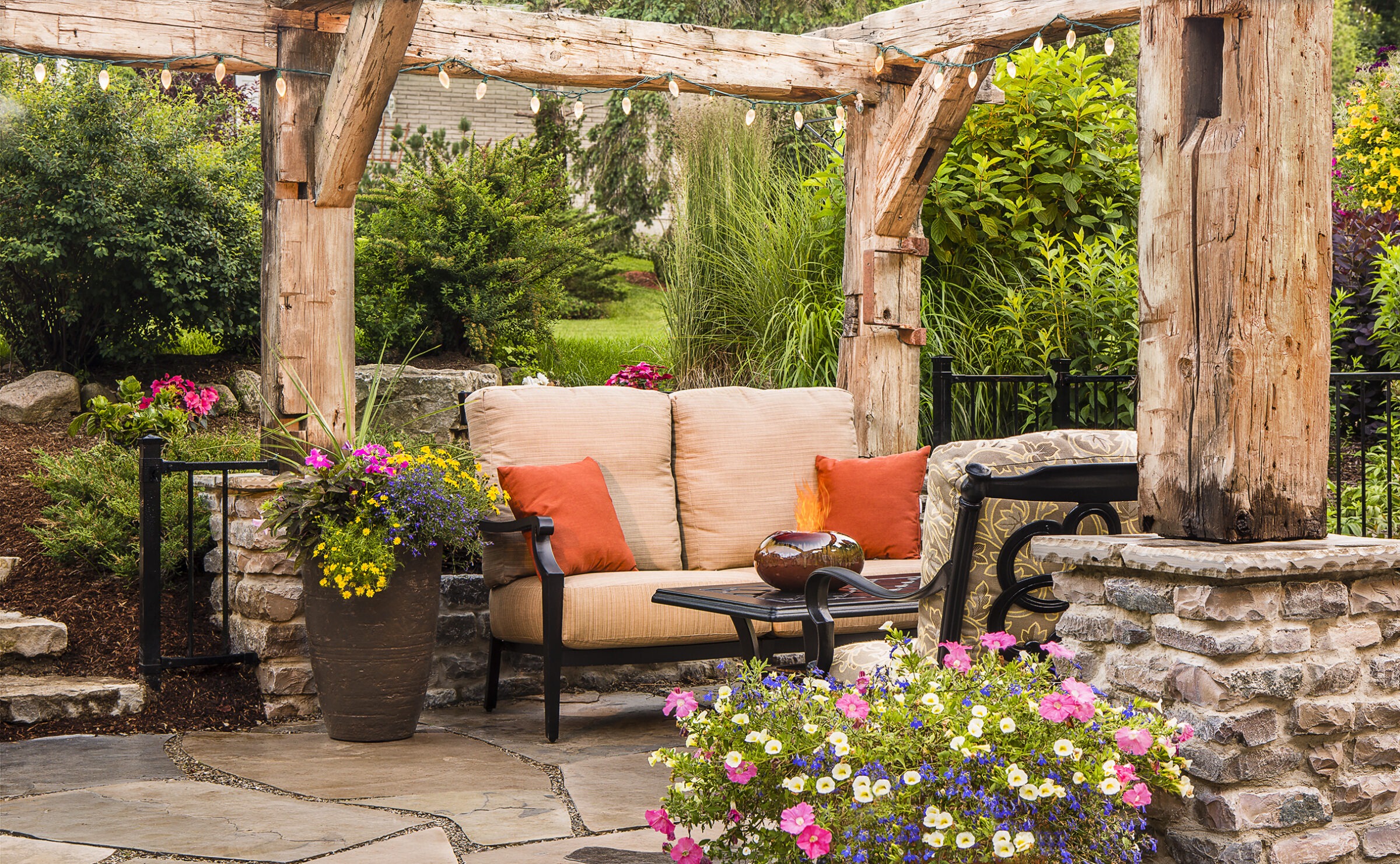 An inviting outdoor patio area with rustic wooden pergola, comfortable seating, colorful flowers, stone walls, and decorative string lights.