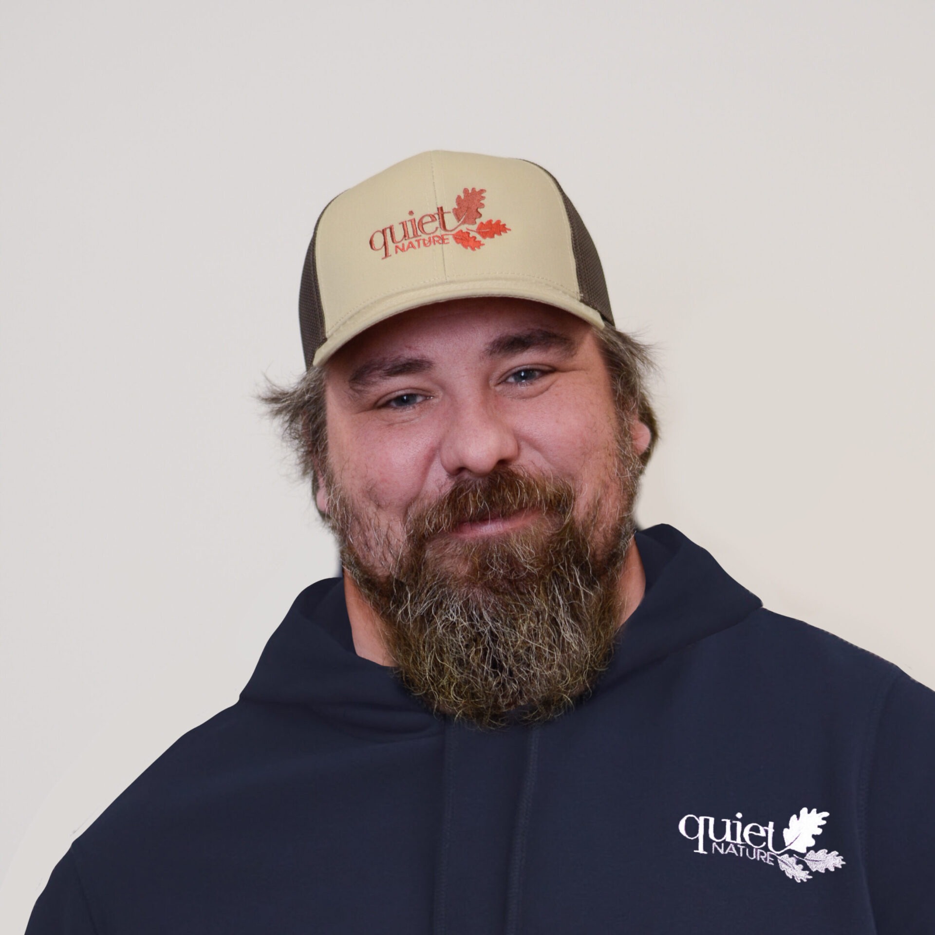A person with a full beard is smiling at the camera, wearing a baseball cap and a hoodie with the "Quiet Nature" logo.
