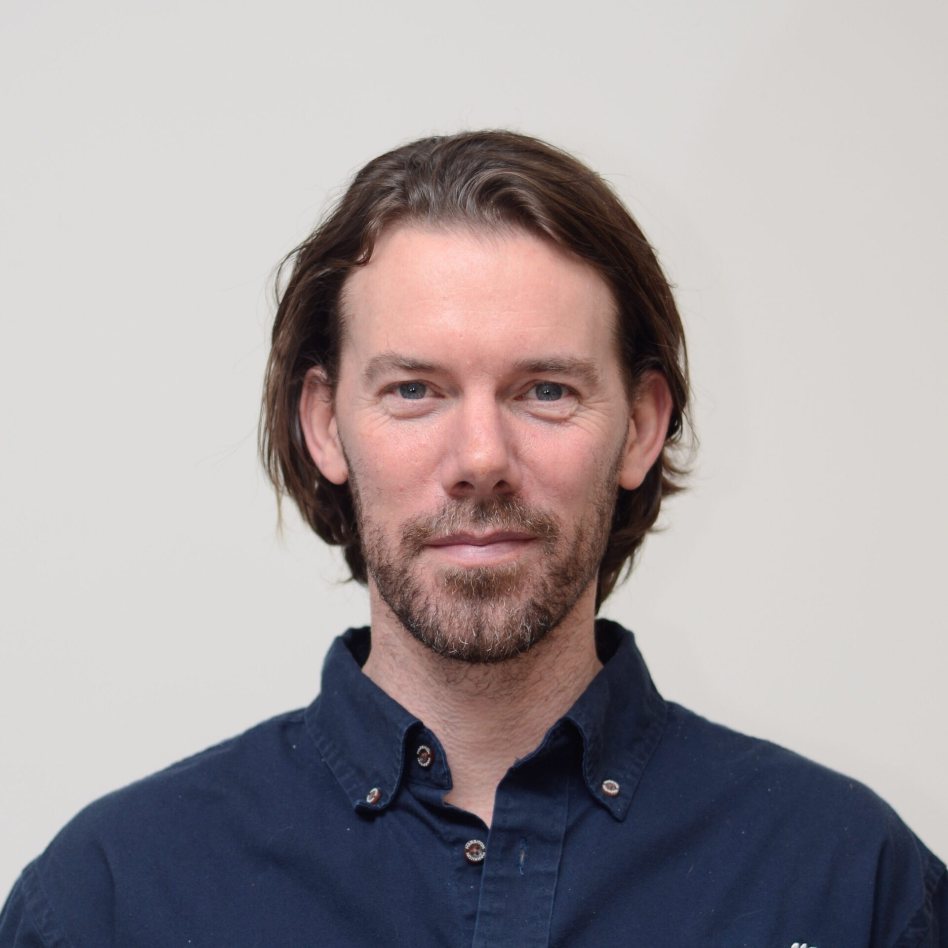 The image shows a person with medium-length brown hair, blue eyes, slight stubble, wearing a dark shirt, standing against a plain background.