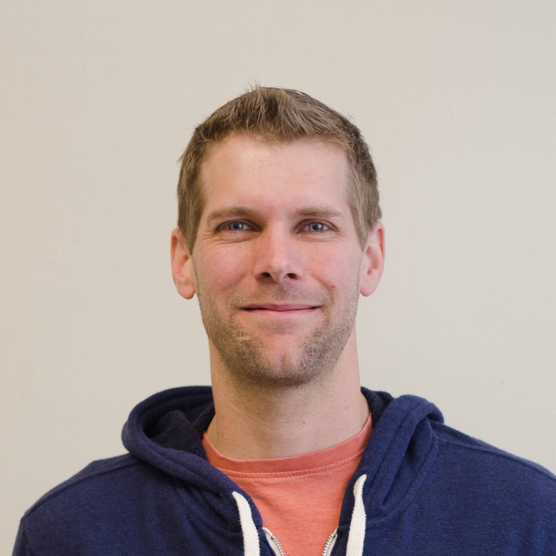 A smiling person with short hair is wearing a blue hoodie over a coral shirt, against a pale background. They exhibit a friendly demeanor.