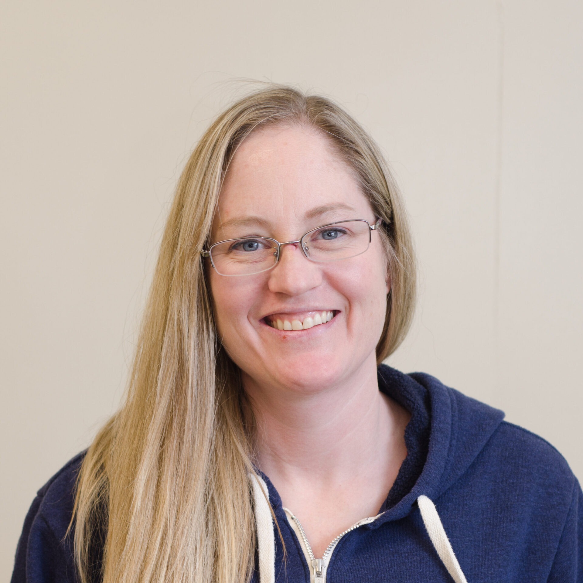 A smiling person with long blonde hair and glasses, wearing a blue hoodie, is against a plain beige background, looking directly at the camera.