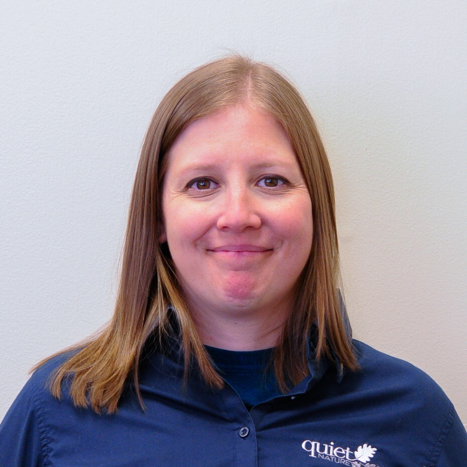 A person with straight shoulder-length hair, smiling lightly at the camera, wearing a dark blue collared shirt with a "Quiet Nature" logo.