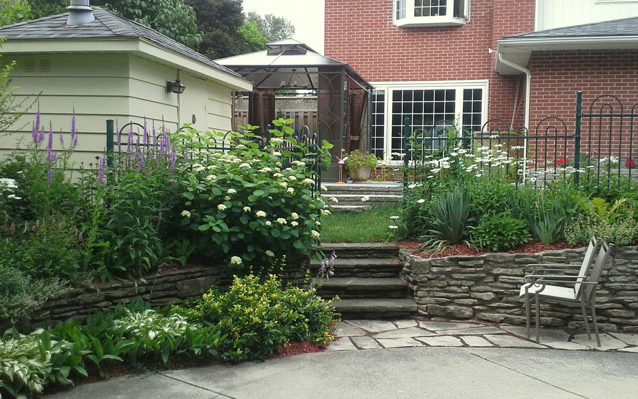 This image shows a well-maintained garden with a stone pathway and steps, colorful flowers, and a gazebo beside a red brick house with white windows.