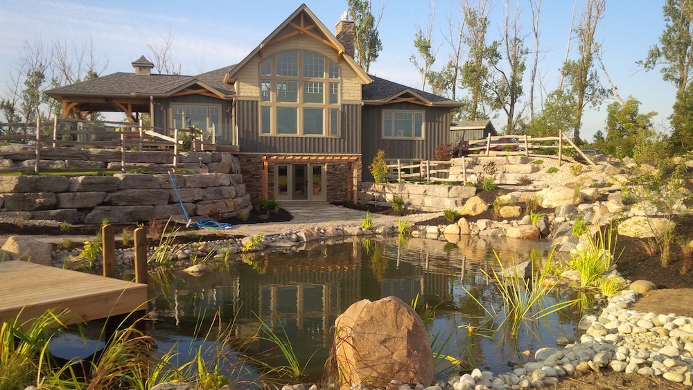 A rustic house with large windows and a wooden deck overlooks a landscaped pond with rocks, plants, and a wooden dock on a sunny day.