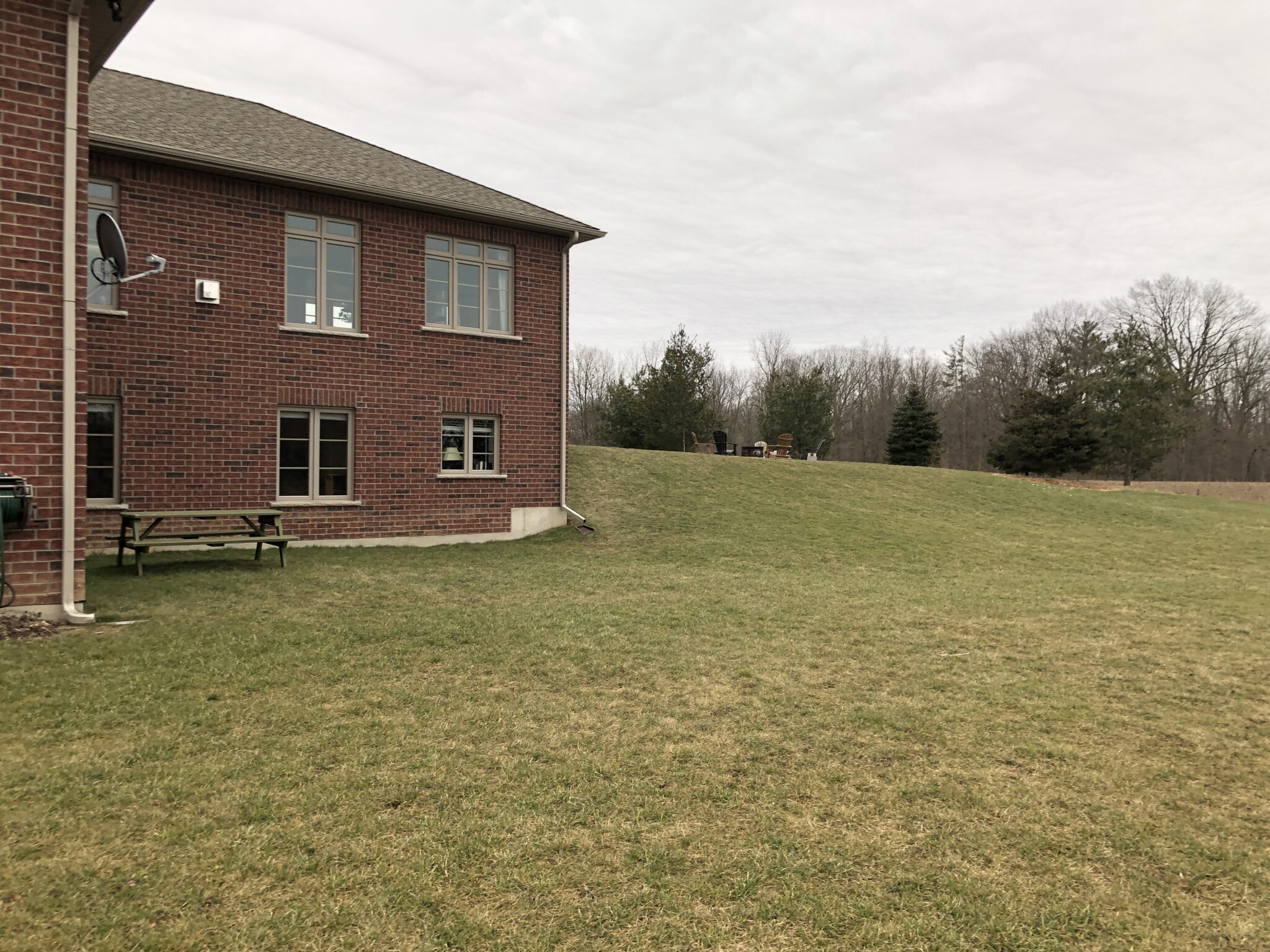 A two-story brick house with multiple windows and a satellite dish. A green lawn slopes upward towards trees and an overcast sky.
