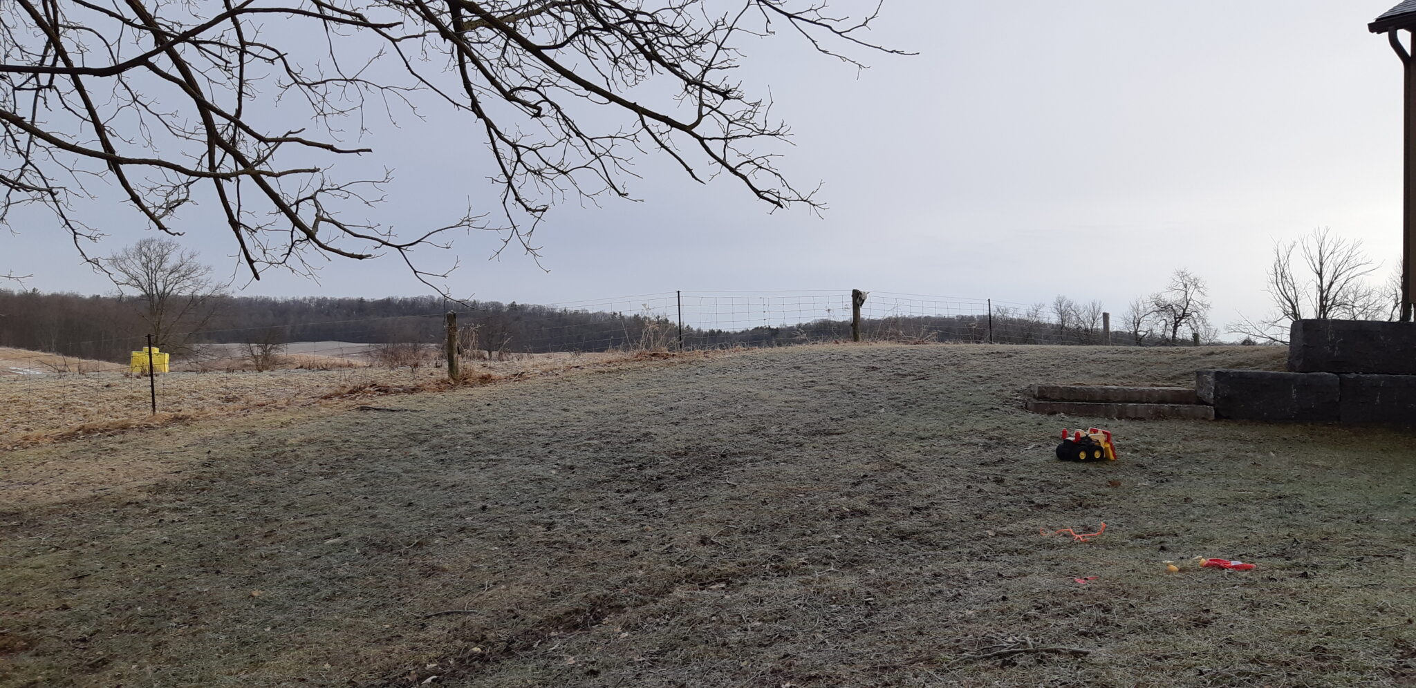A frosty rural landscape with bare trees, a wire fence, children's toys on the ground, a water body in the distance, and overcast skies.