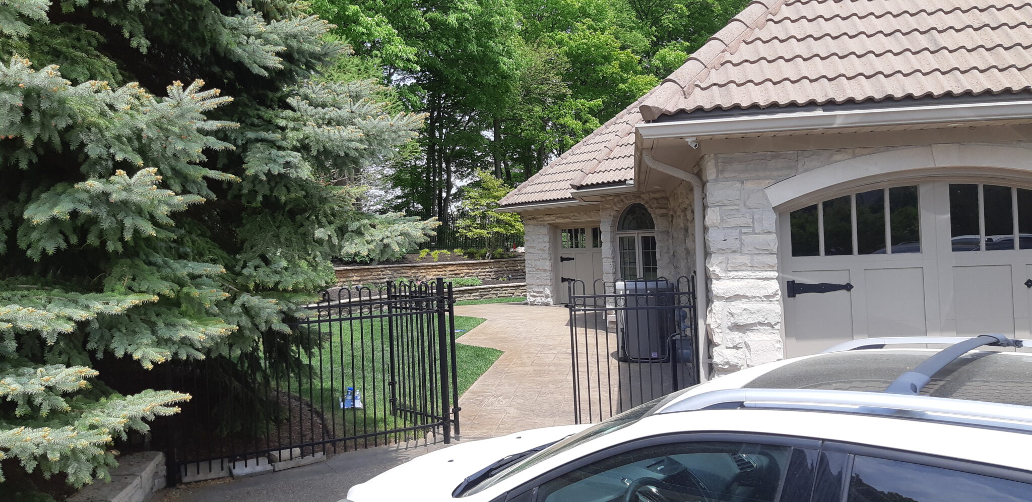 A stone house with a double garage door, flanked by evergreen trees and a wrought-iron gate, on a sunny day with clear skies. Partial view of a car.