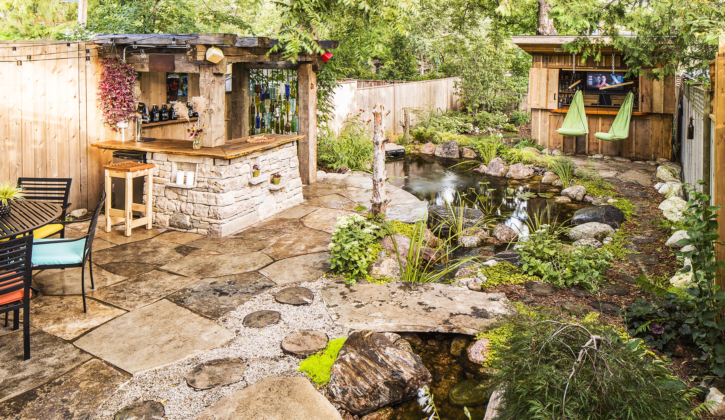 An outdoor space features a bar, hanging seats, a TV cabinet, stone paths, and a lush pond surrounded by vegetation and wooden fences.