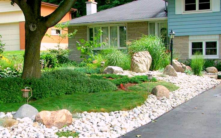 A neatly landscaped front yard with lush greenery, large rocks, a pebble path, and a traditional house with a visible street lamp post.