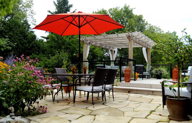 An inviting outdoor patio with a bright red umbrella, dining chairs, a pergola, and lush surrounding greenery and blooming flowers.