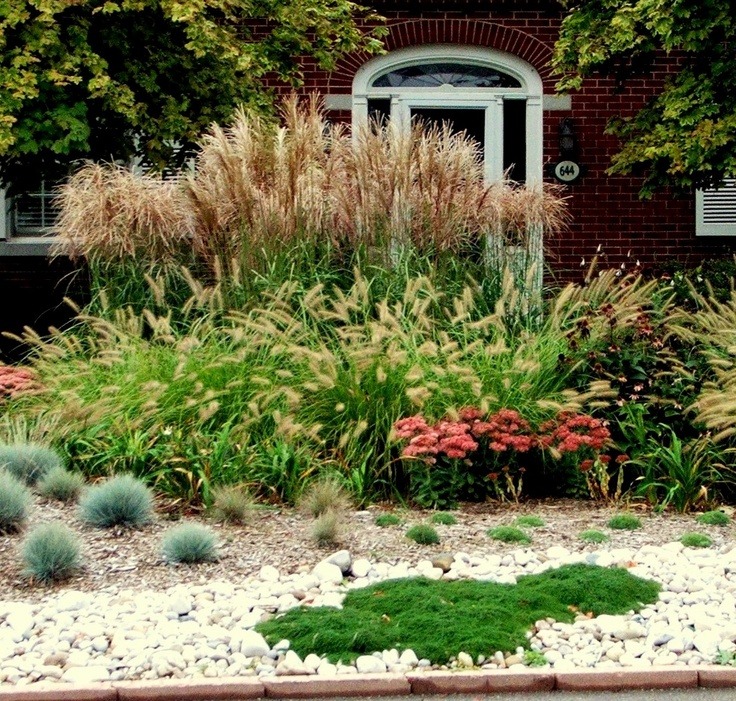 A landscaped front yard with ornamental grasses, flowering plants, and decorative stones in front of a red brick house with a white arched window.