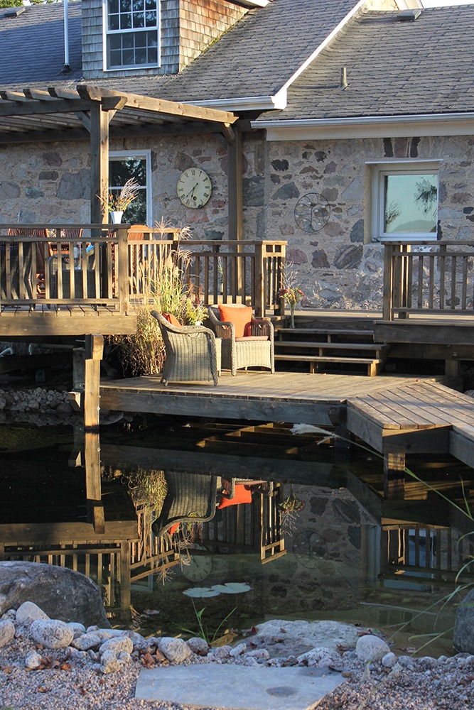 A rustic stone house with a wooden deck, wicker chairs, and decorative plants. A clock on the wall and a calm pond reflect the scene.