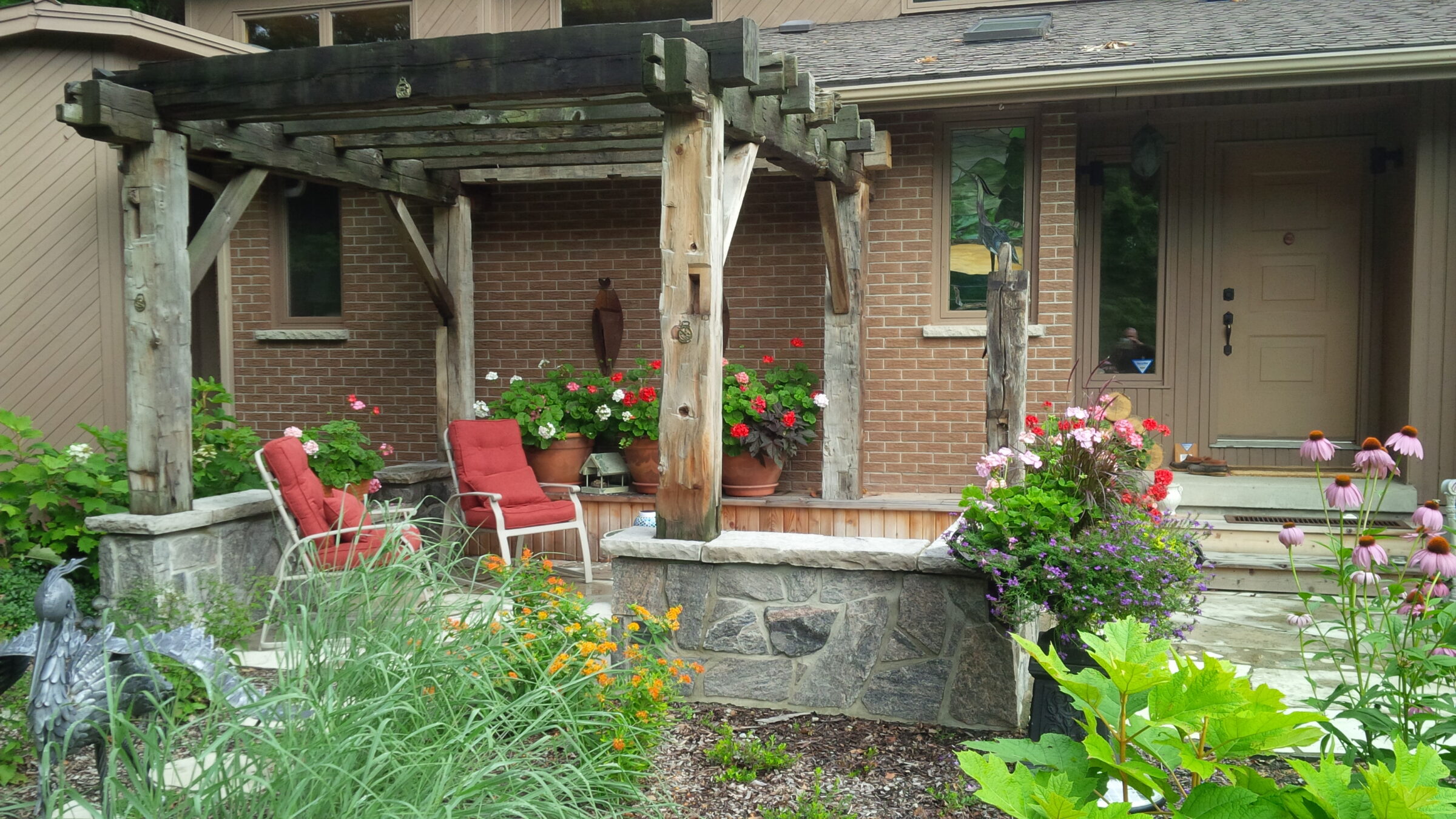 A peaceful home entrance with a wooden pergola, red cushioned chairs, potted flowers, decorative sculptures, and blooming garden plants.
