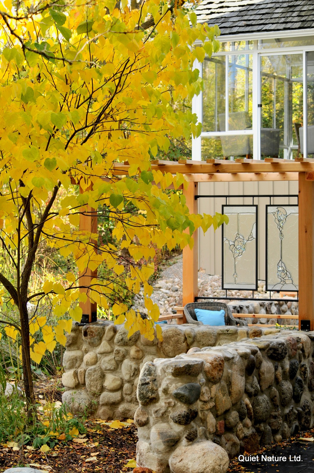 A serene autumn setting featuring a stone wall, yellow foliage, and a part of a house with decorative glass panels and wooden elements.