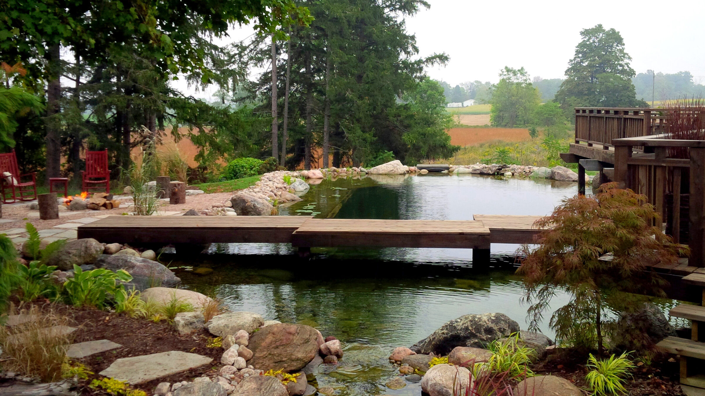 A serene outdoor setting with a wooden deck stretching over a tranquil pond, surrounded by rocks, trees, and red chairs, with a distant field visible.