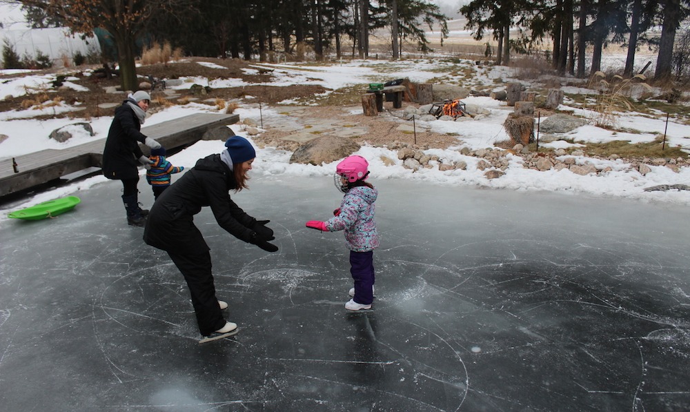 Three individuals are ice skating on a frozen pond. Two adults assist a child. There's a campfire, green sled, and melting snow in the background.