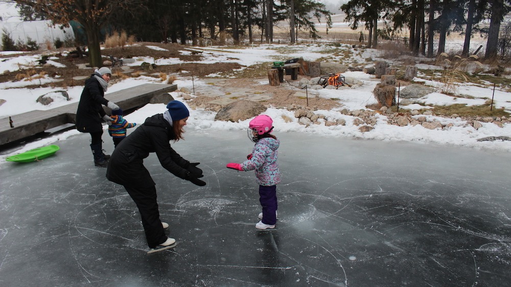 A person teaches a child to ice skate while another adult and child watch nearby. They're on a homemade rink with snow and a fire pit visible.